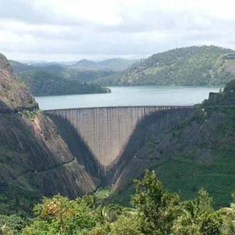 A large concrete dam spanning a valley with water behind it, surrounded by forested hills under a cloudy sky.