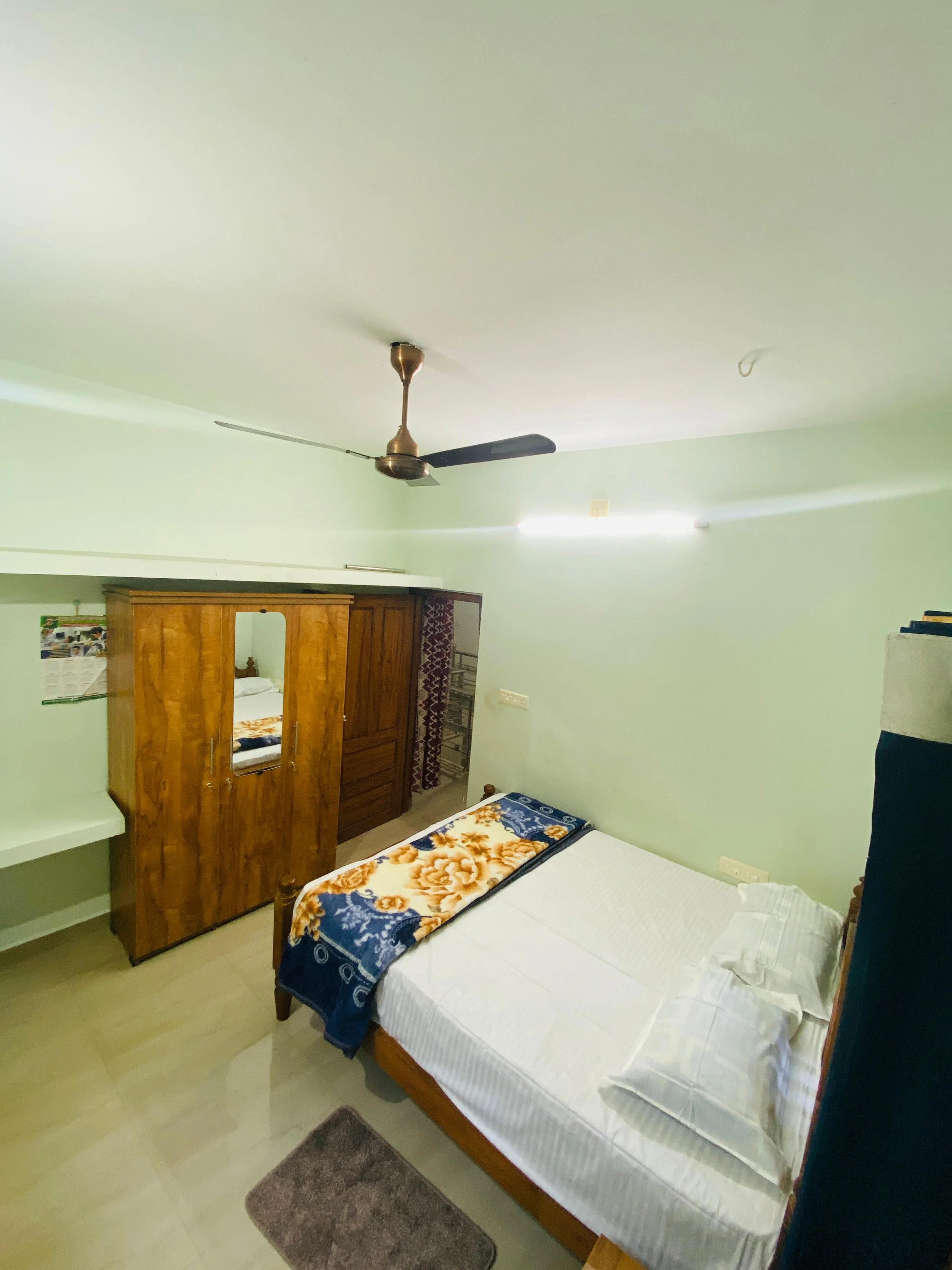 A bedroom with a wooden wardrobe, a bed with white bedding and a blue and yellow blanket, a ceiling fan, a small gray rug, and a window with dark curtains.