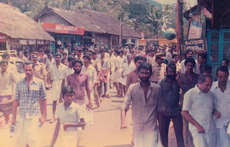 Crowd of men walking on a street in a rural area with small shops and traditional houses.