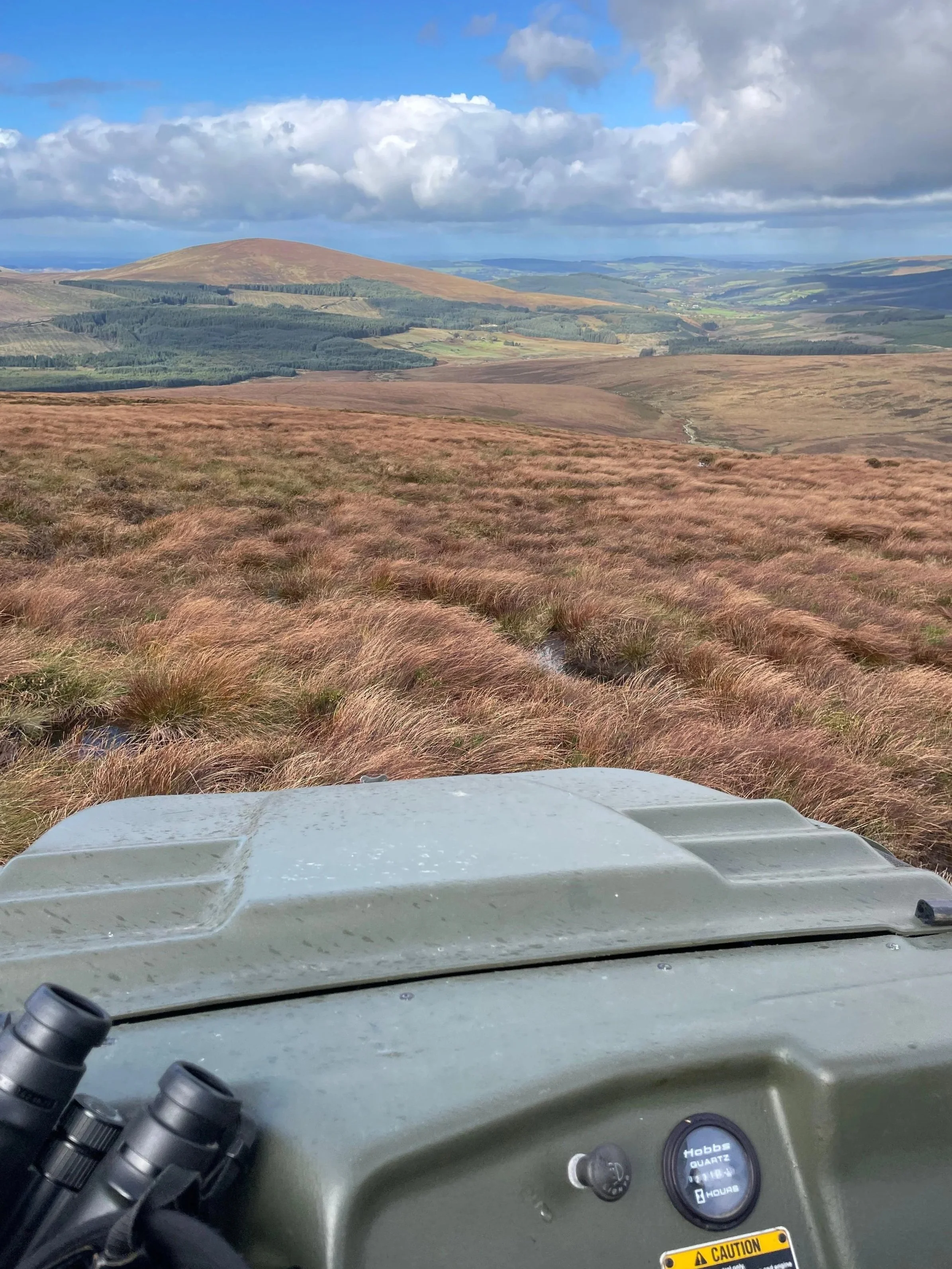 View from a vehicle showing a landscape with rolling hills covered in brown grass, a cloudy sky, and distant green fields and patches of forest.