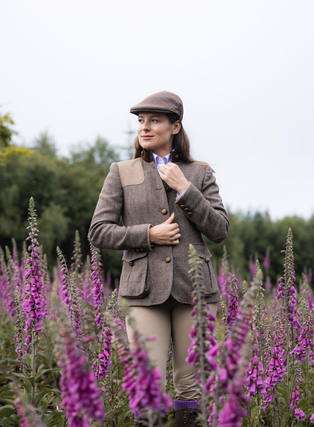 Woman in vintage hunting attire standing among pink foxglove flowers in a field with trees in the background.
