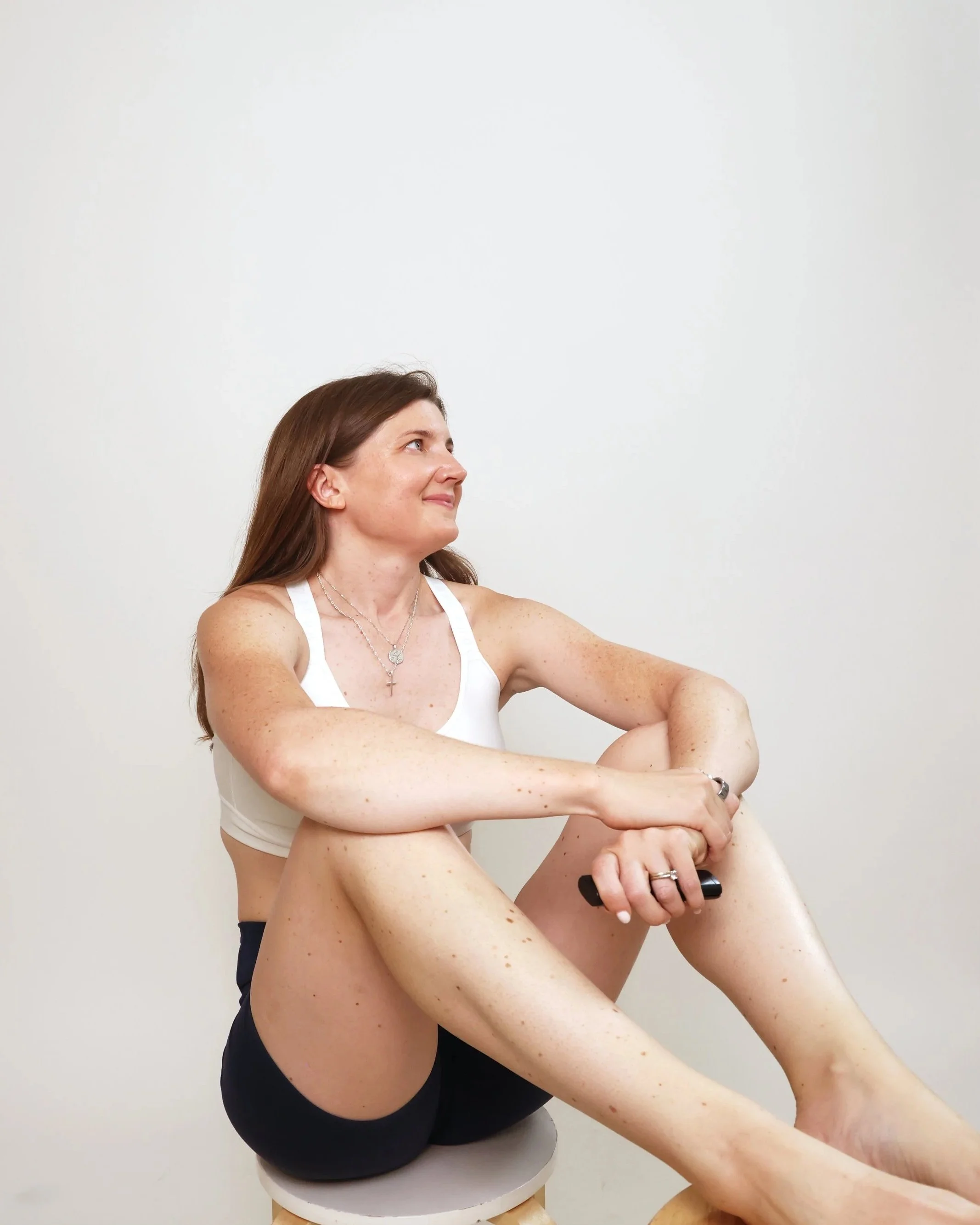 Woman with long brown hair sitting on a stool against a plain white wall, smiling, holding a black object, wearing a white sports bra, black shorts, and layered necklaces.