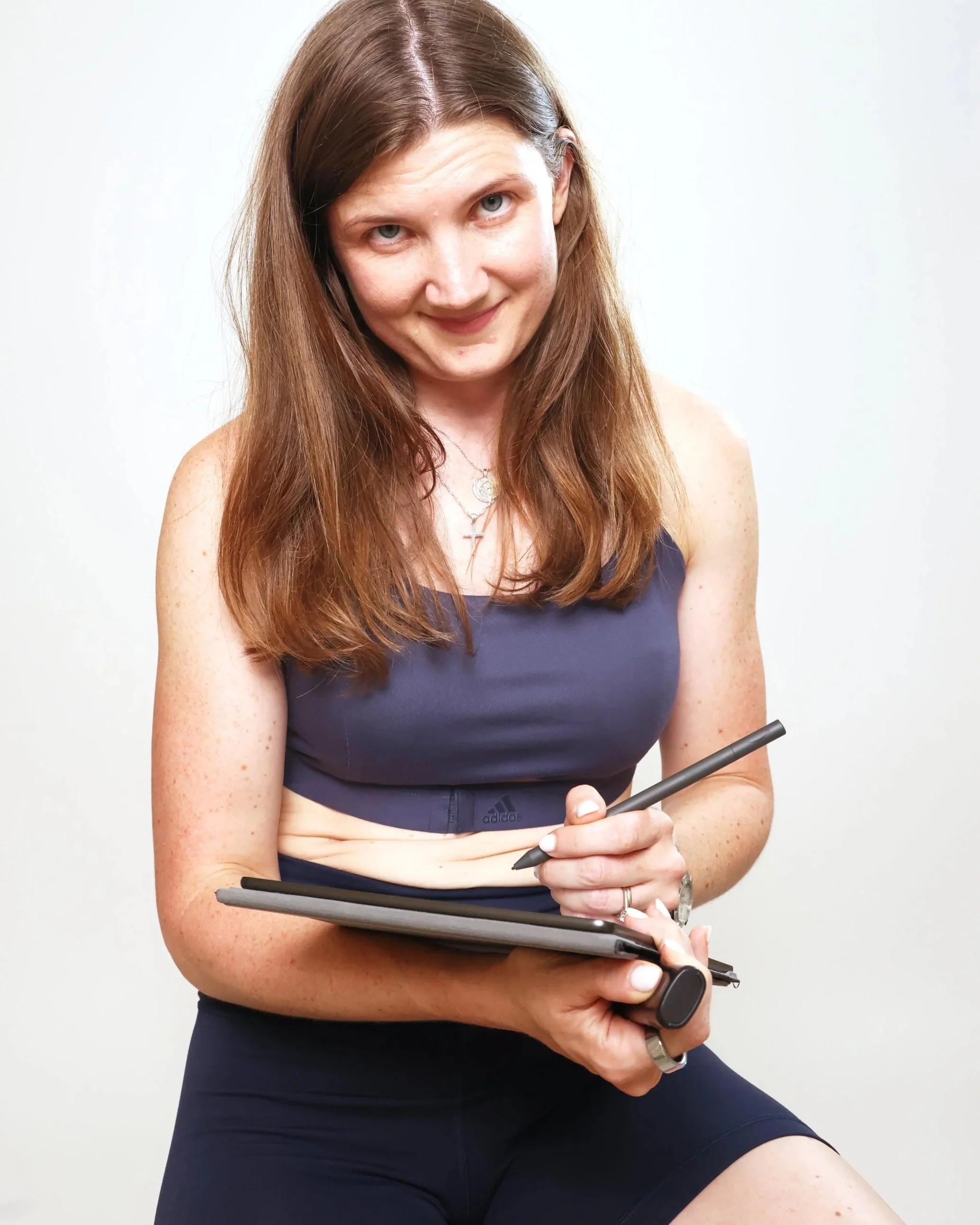 A woman with long brown hair, wearing a navy sports bra and black shorts, holding a stylus and a digital drawing tablet, smiling at the camera.