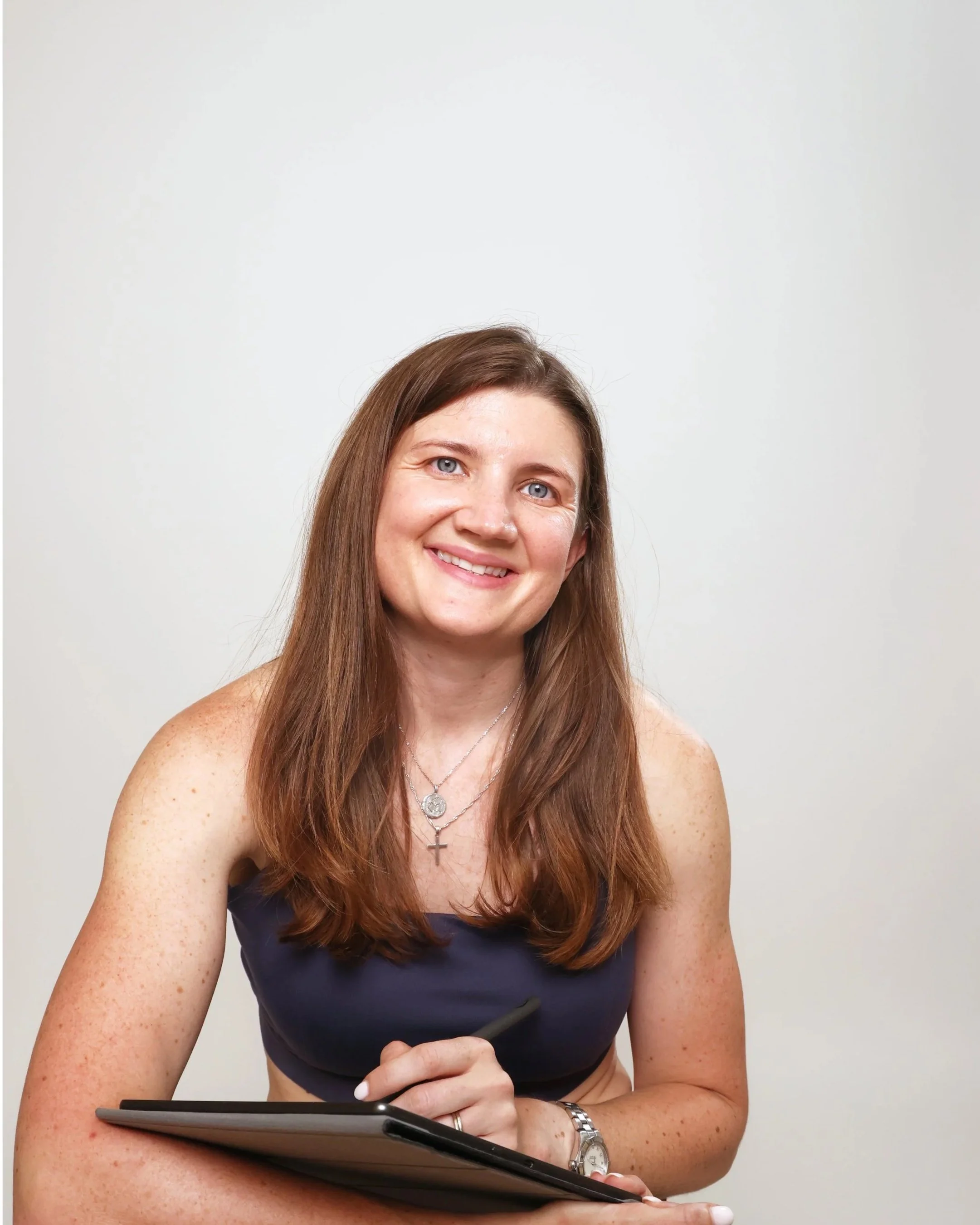 A woman with long reddish-brown hair smiling, holding a digital pen and a tablet against a plain light gray background.