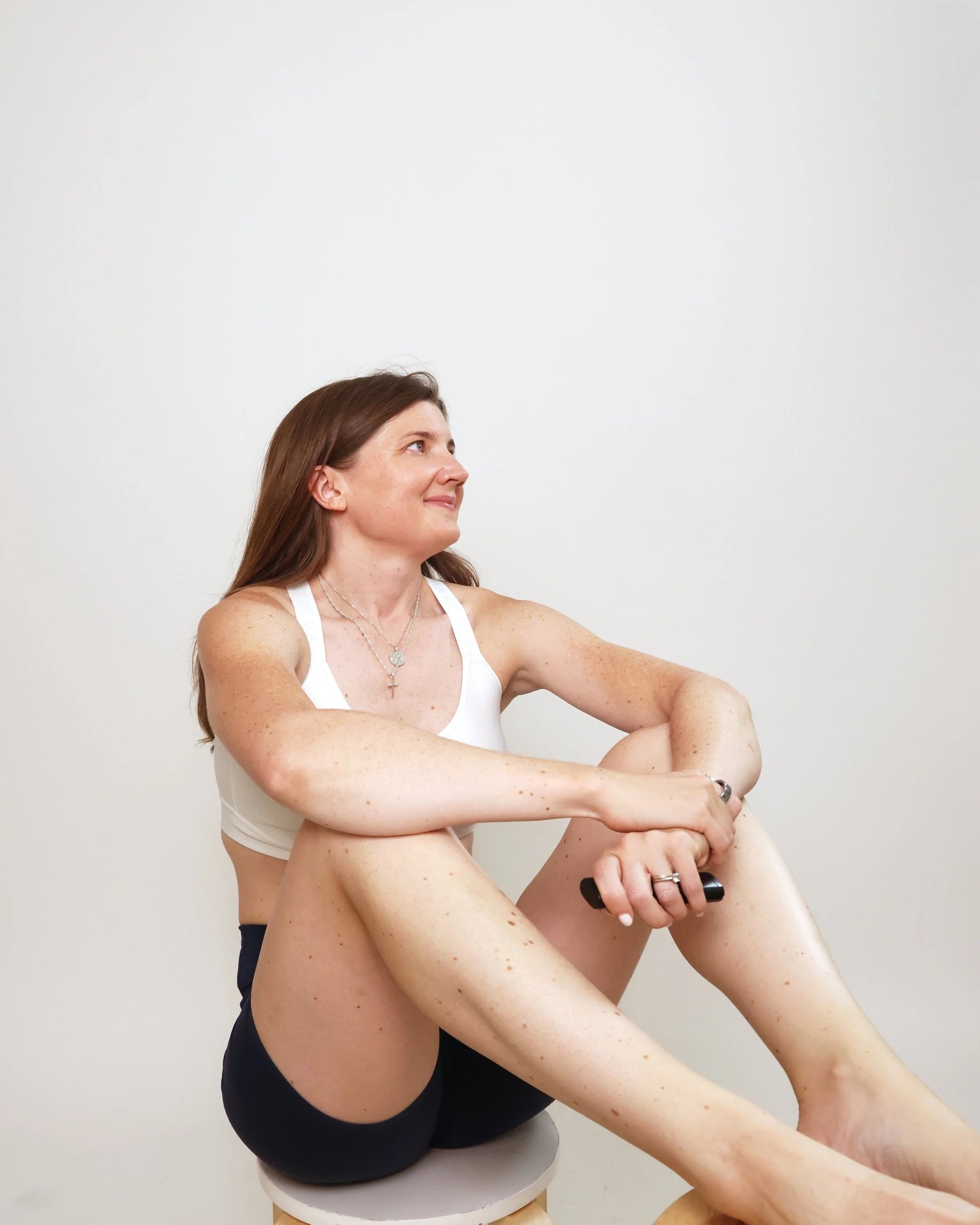 Woman with brown hair wearing a white sports bra and black shorts sitting on a stool against a plain white wall, smiling and holding a black object.