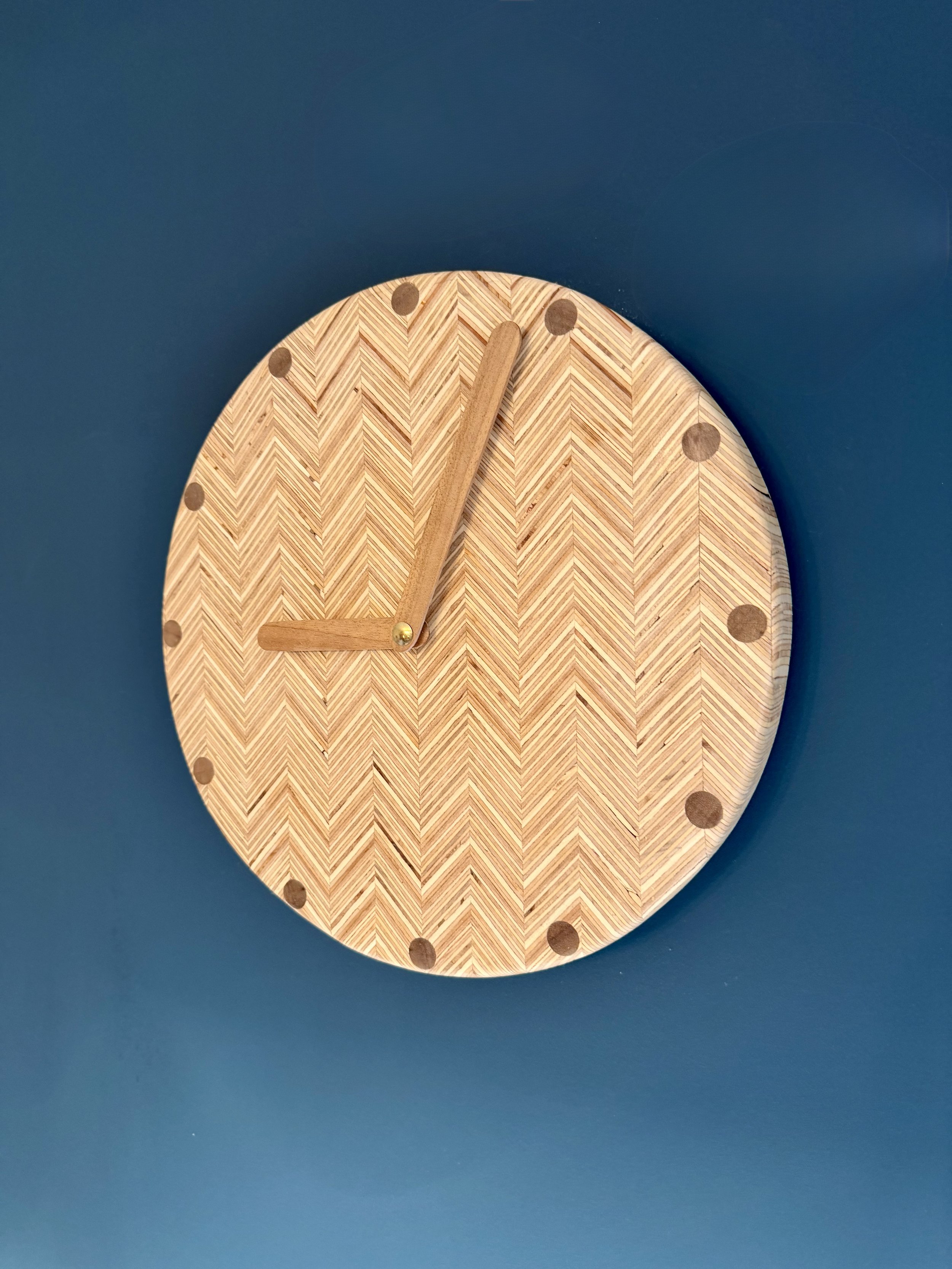 A round wooden wall clock with a chevron pattern, dark dots at each hour mark, and wooden hands showing the time as 3:00, mounted on a blue wall.