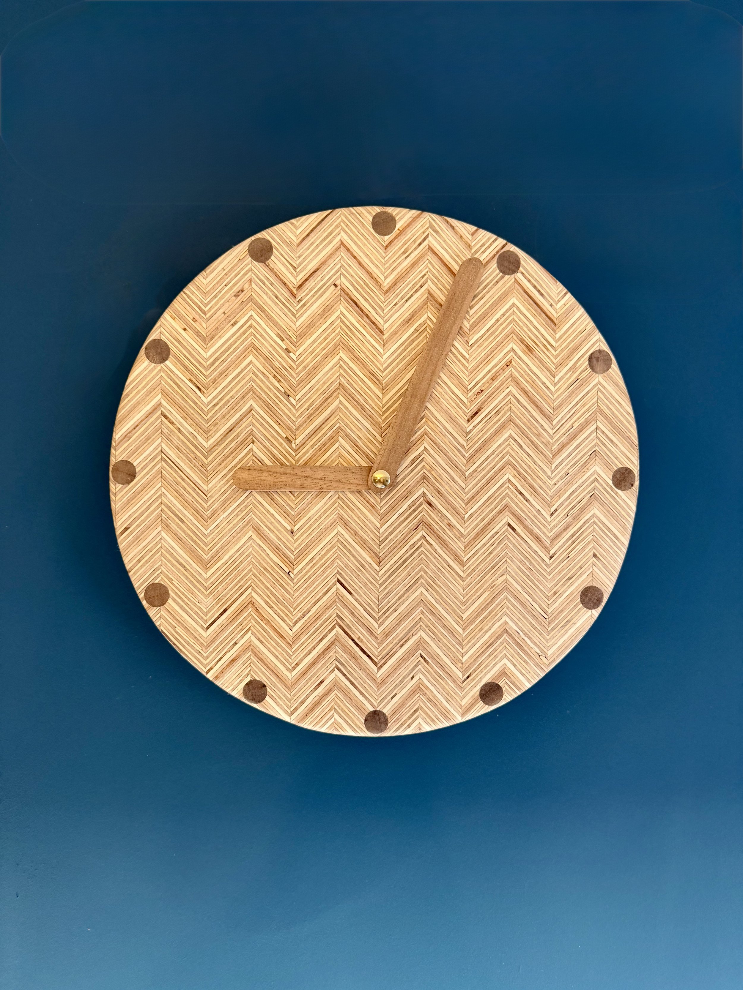 Wooden wall clock with a herringbone pattern, black dot markers, and simple hour and minute hands, showing the time as 3:05.