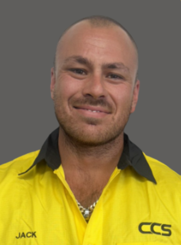 Portrait of a young man with a shaved head, wearing a yellow work shirt with black accents, a chain necklace, and a slight smile, against a gray background.
