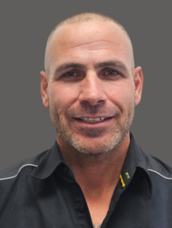 Close-up of a smiling man with a shaved head, stubble, wearing a black collared shirt against a gray background.