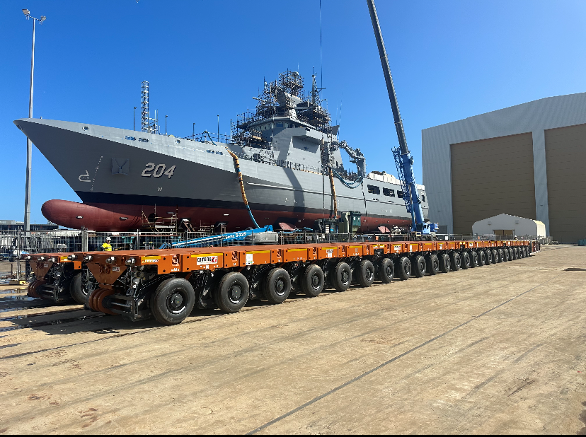 A large naval ship with the number 204 on its hull, being transported on an extensive orange flatbed trailer with multiple wheels, next to a warehouse-like building, under a clear blue sky.