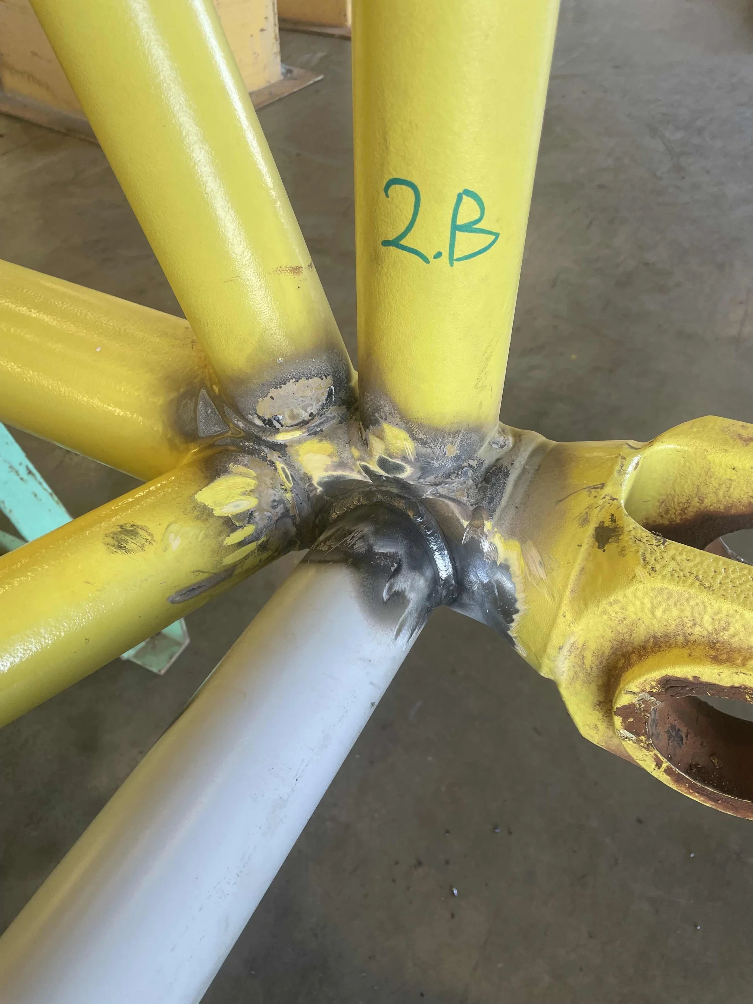 Close-up of a yellow metal structure with welded joints and the marking '2B' written in blue ink.