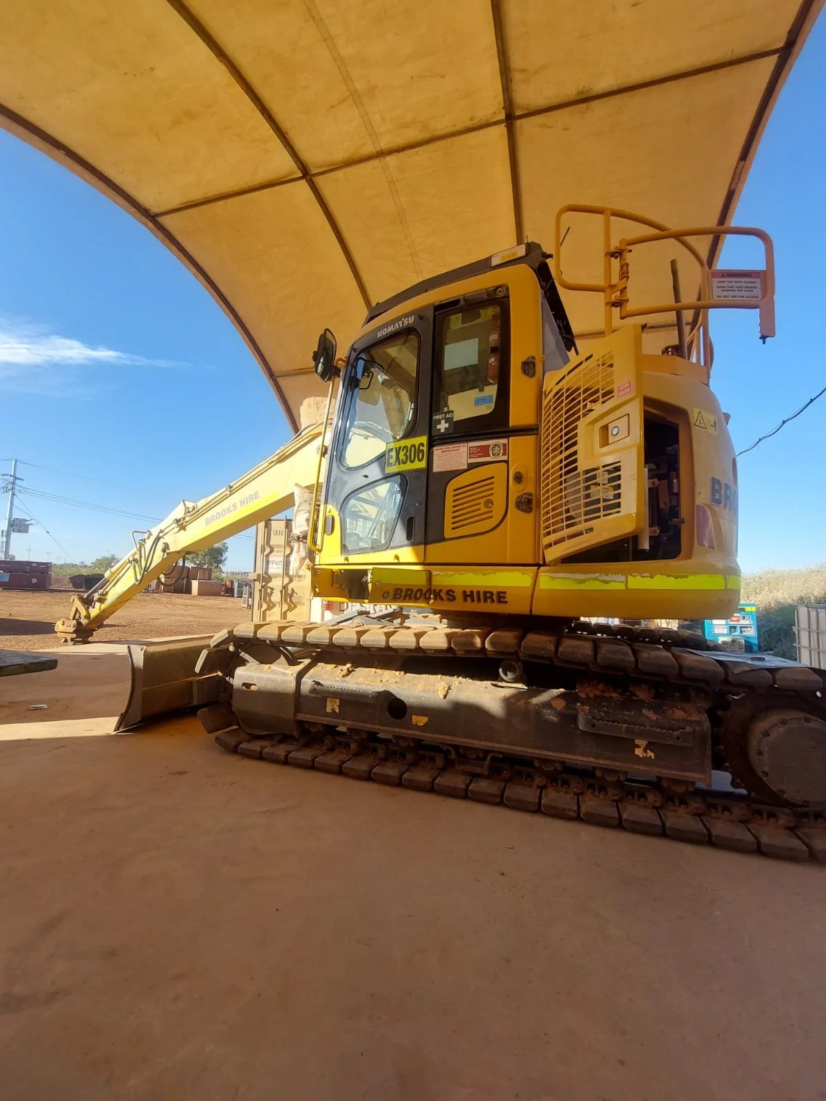 A yellow construction excavator with tracks, parked under a large, curved canopy, with a blue sky and some distant vehicles in the background.