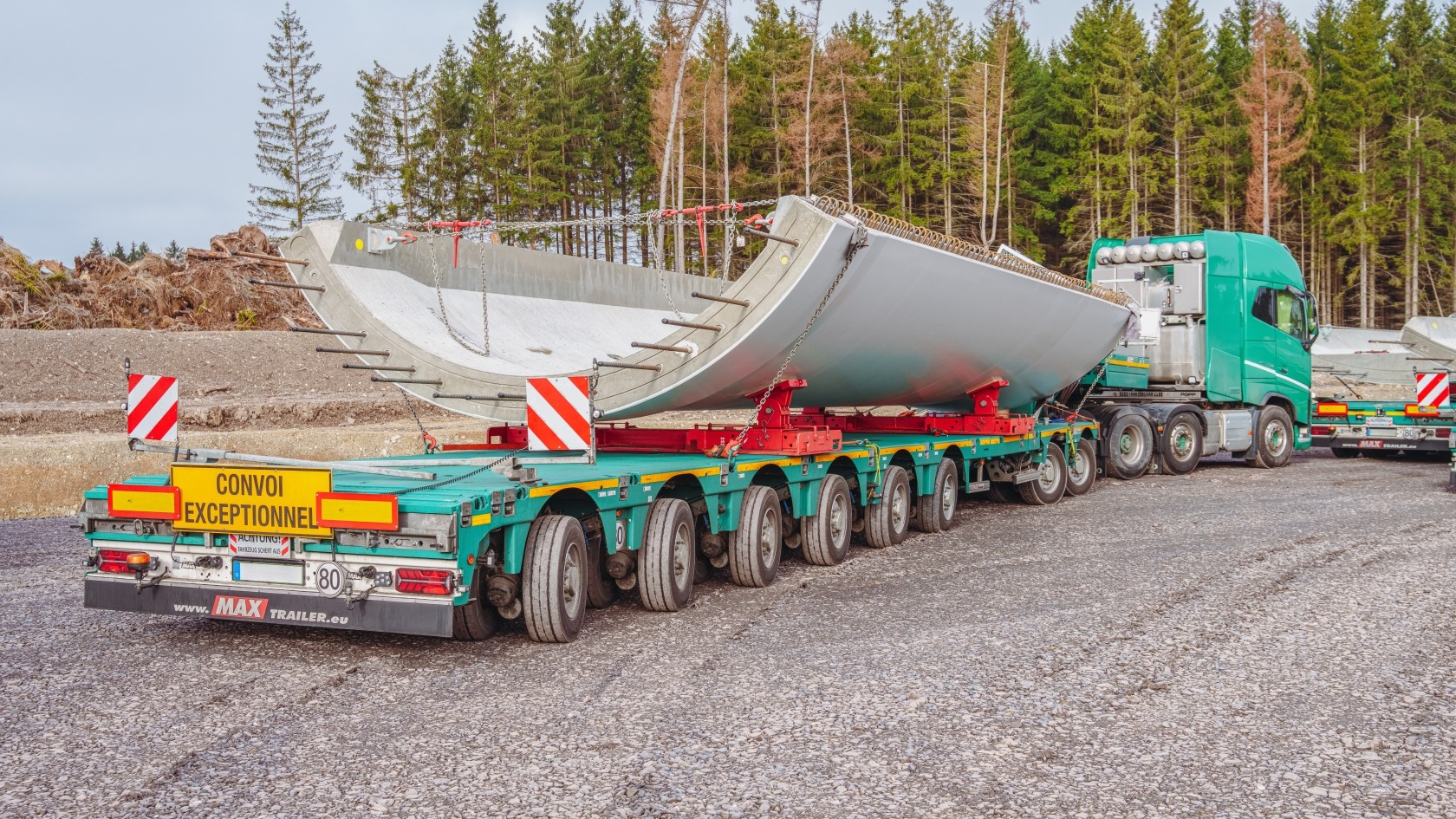 Large green semi-truck transporting a massive ship propeller on a trailer, with a small yellow sign that reads 'CONVOI EXCEPTIONNEL' on the back and red and white caution signs. The background features a forest of tall pine trees.