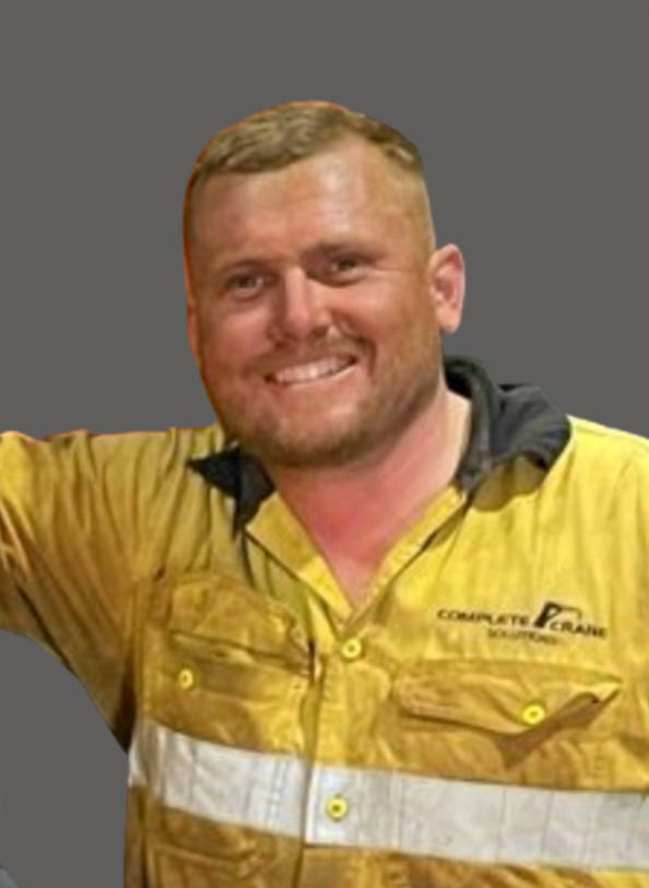 Man with light brown hair and beard smiling wearing a yellow and black work shirt with patches, standing against a gray background.