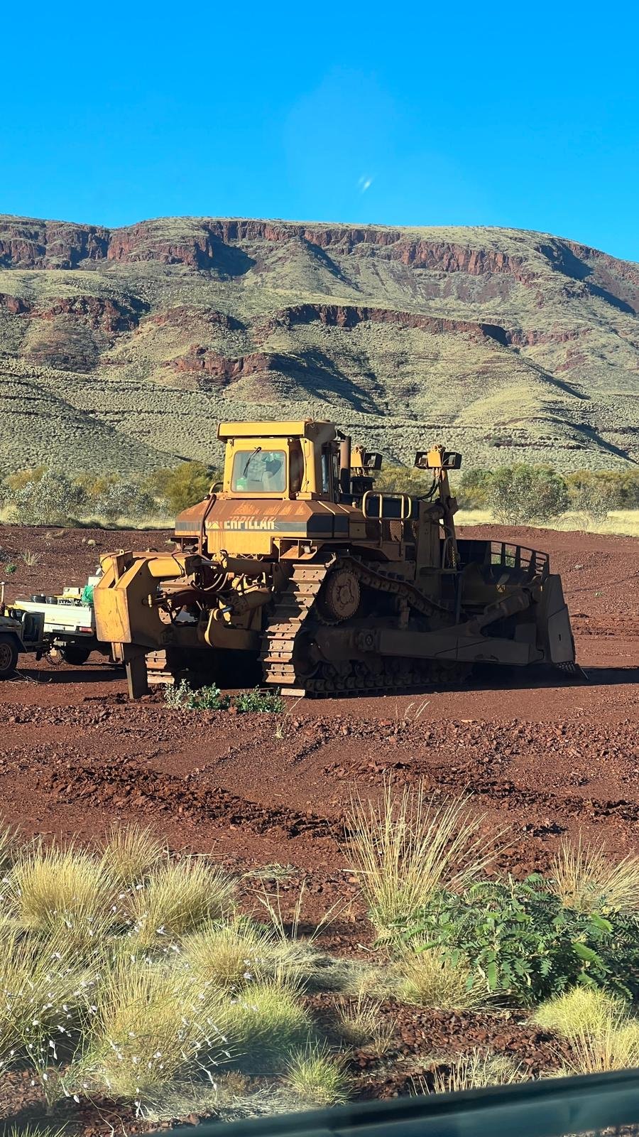 A bulldozer on a dirt path with desert plants in the foreground and mountains in the background under a clear blue sky.