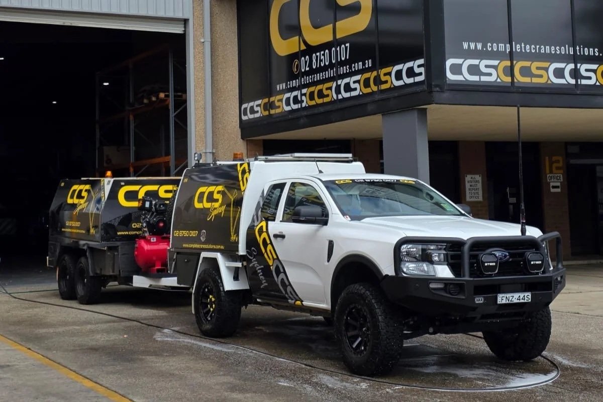 A white utility truck with black wheels and a black front bumper guard parked outside a building with signage for CCS Solutions. The truck is towing a utility trailer with branding matching the company's colors and logo.