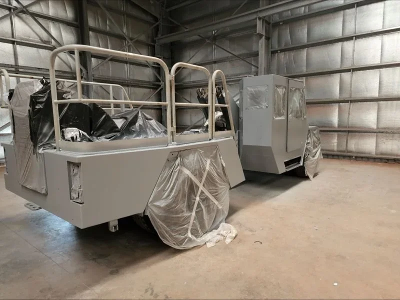 Two large, rectangular industrial vehicles in a warehouse, covered partially with plastic, with some areas masked off, possibly in the process of painting or assembly.