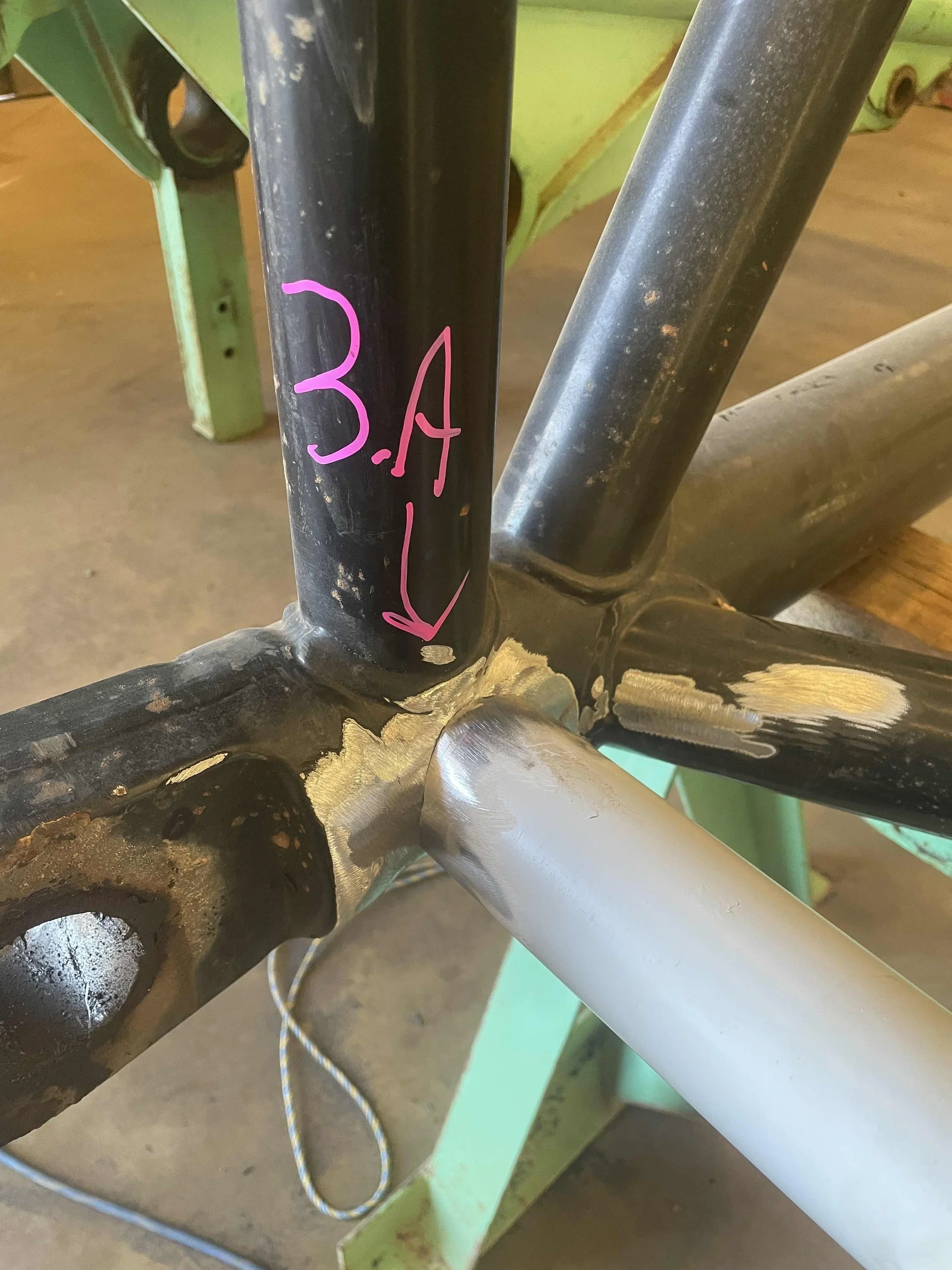 Close-up of a bicycle frame with handwritten pink and red text '3A' pointing to a weld junction, with the bottom part of the bicycle showing signs of rust and wear.