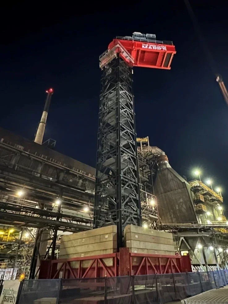 Nighttime view of an oil drill rig with a tall black tower and a red top labeled 'AUGER'. The rig is part of an industrial facility with pipes and structures illuminated by bright lights.