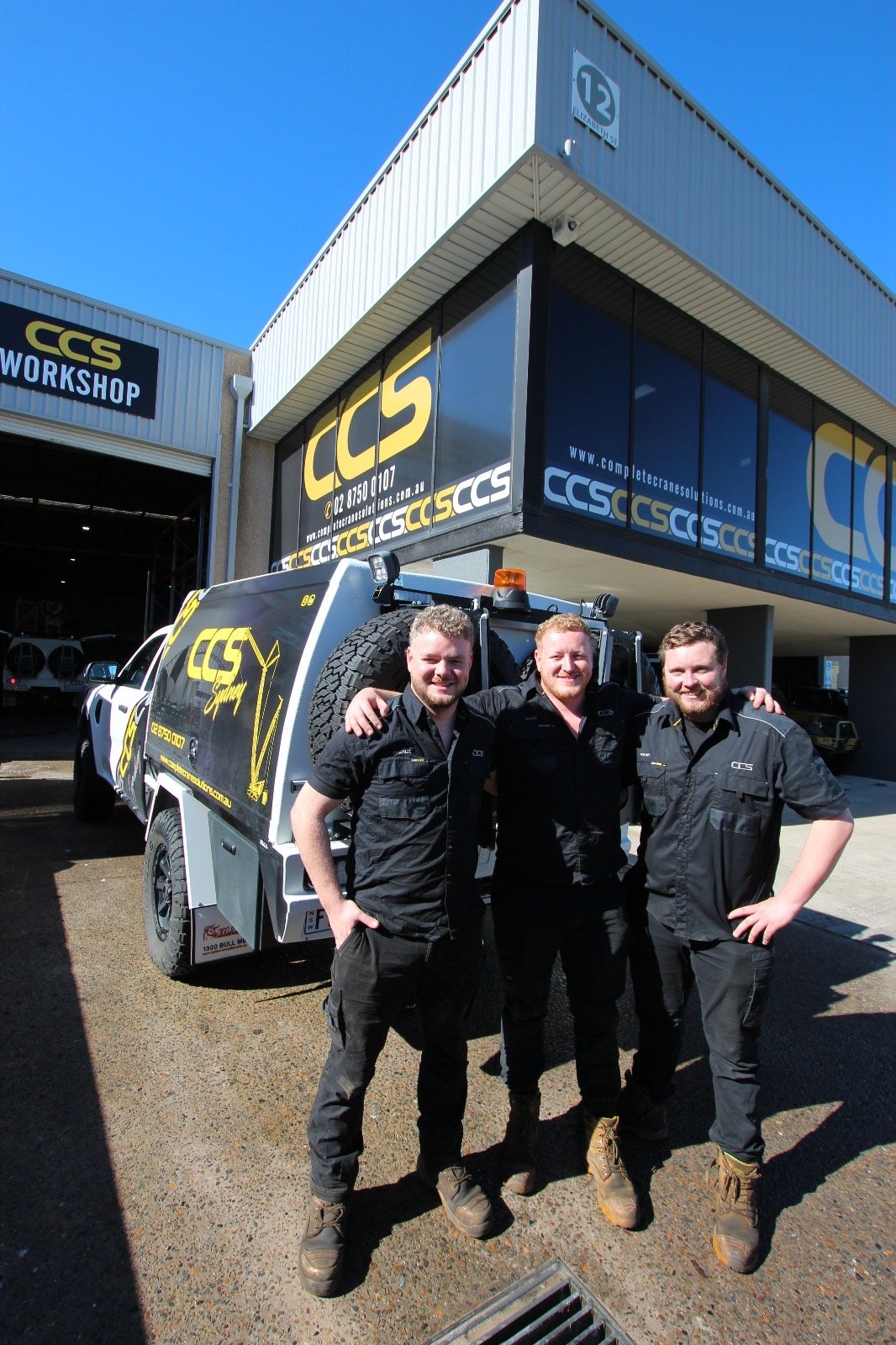 Three smiling men in black uniforms standing outside a workshop with a white vehicle and CCS branding on the building and vehicle.