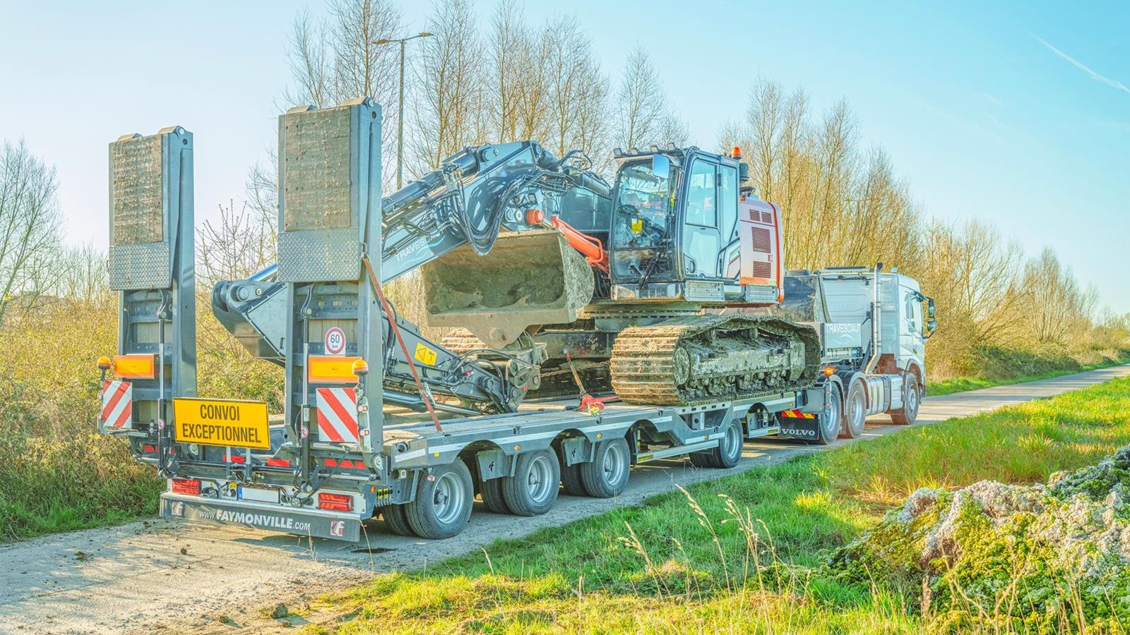 A construction excavator loaded on a flatbed truck on a rural road, with trees and grass in the background.
