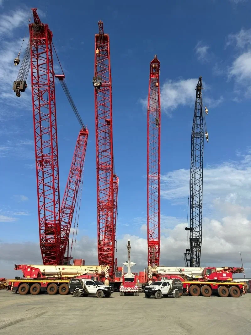 Four large cranes, three painted red and one black, are parked on a flat dirt surface against a blue sky with some clouds. There are two white trucks with extendable arms parked at the base of the cranes.