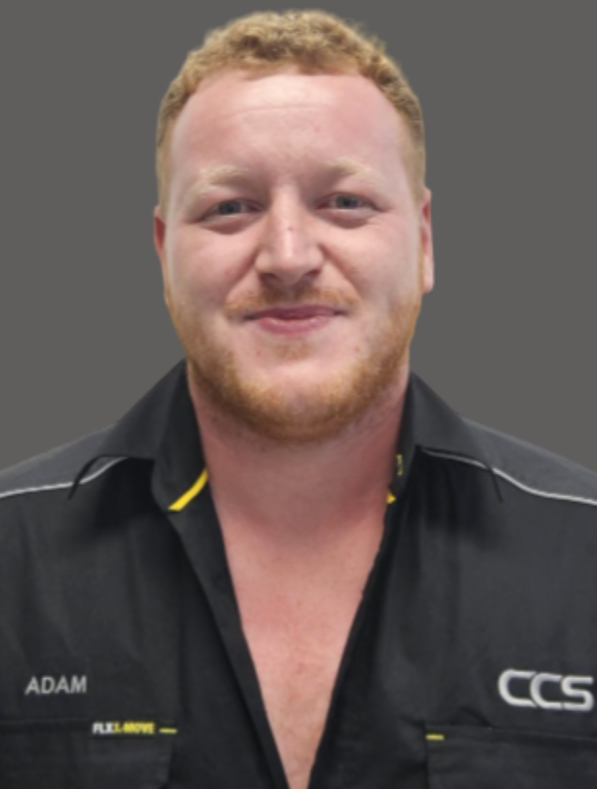 A young man with red hair, beard, and light skin, smiling, wearing a black shirt with yellow accents and the letters 'CCS' on it.