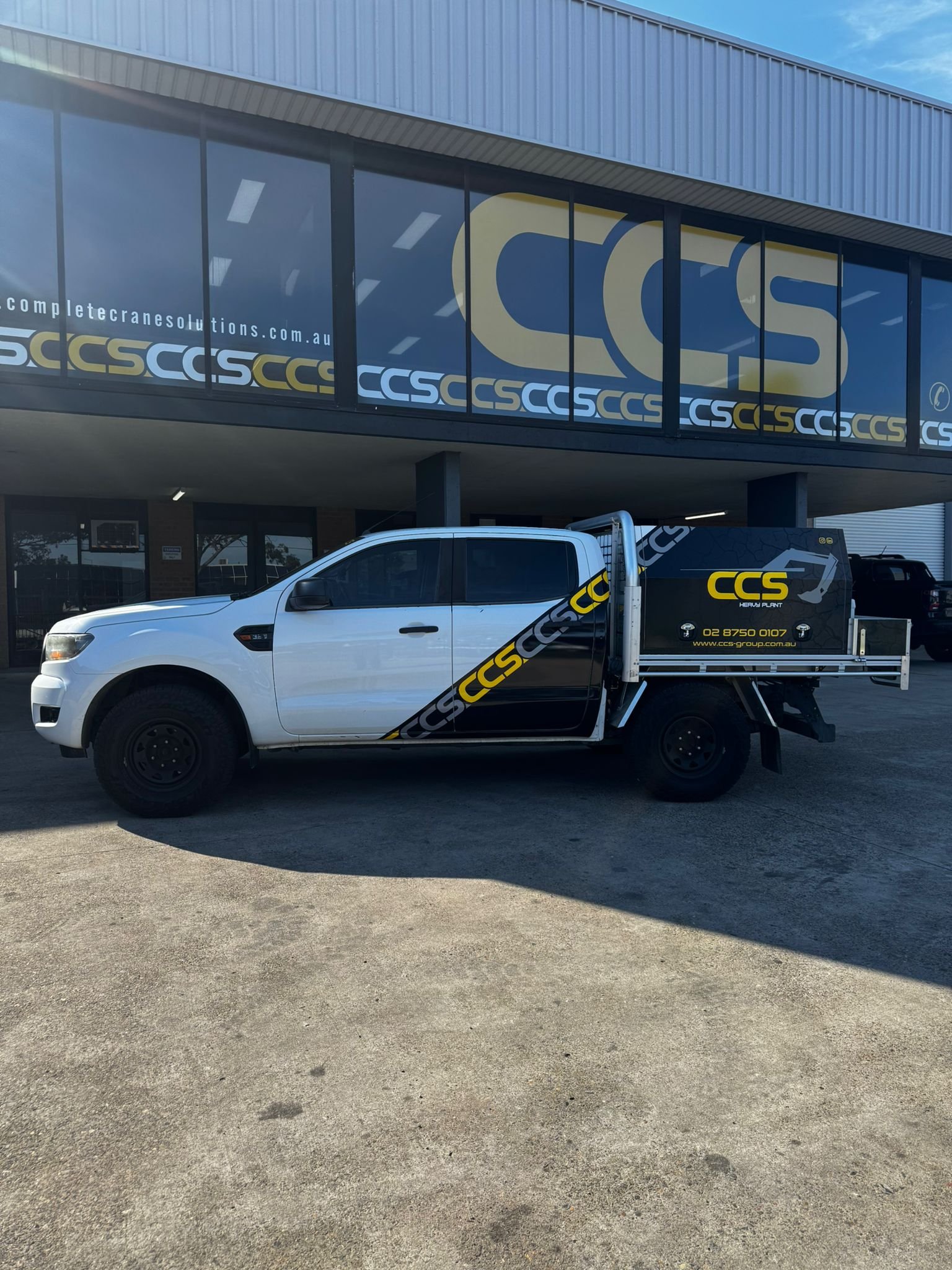 White pickup truck with a black and yellow CCS logo on the door and a black utility bed, parked outside a commercial building with large glass windows displaying the CCS logo and website.