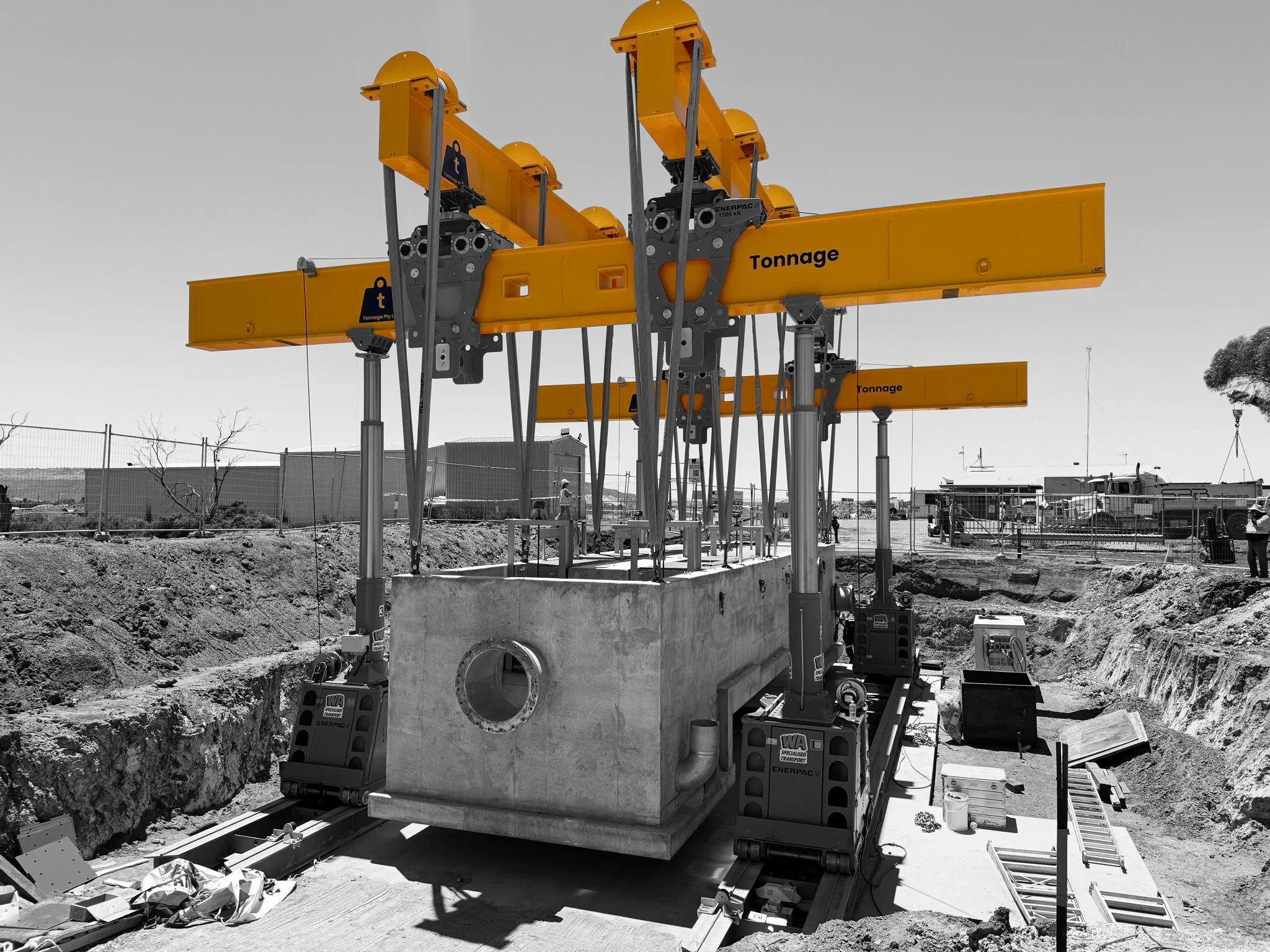 Construction site with large yellow crane lifting concrete section over excavation area with tools and construction materials scattered around.
