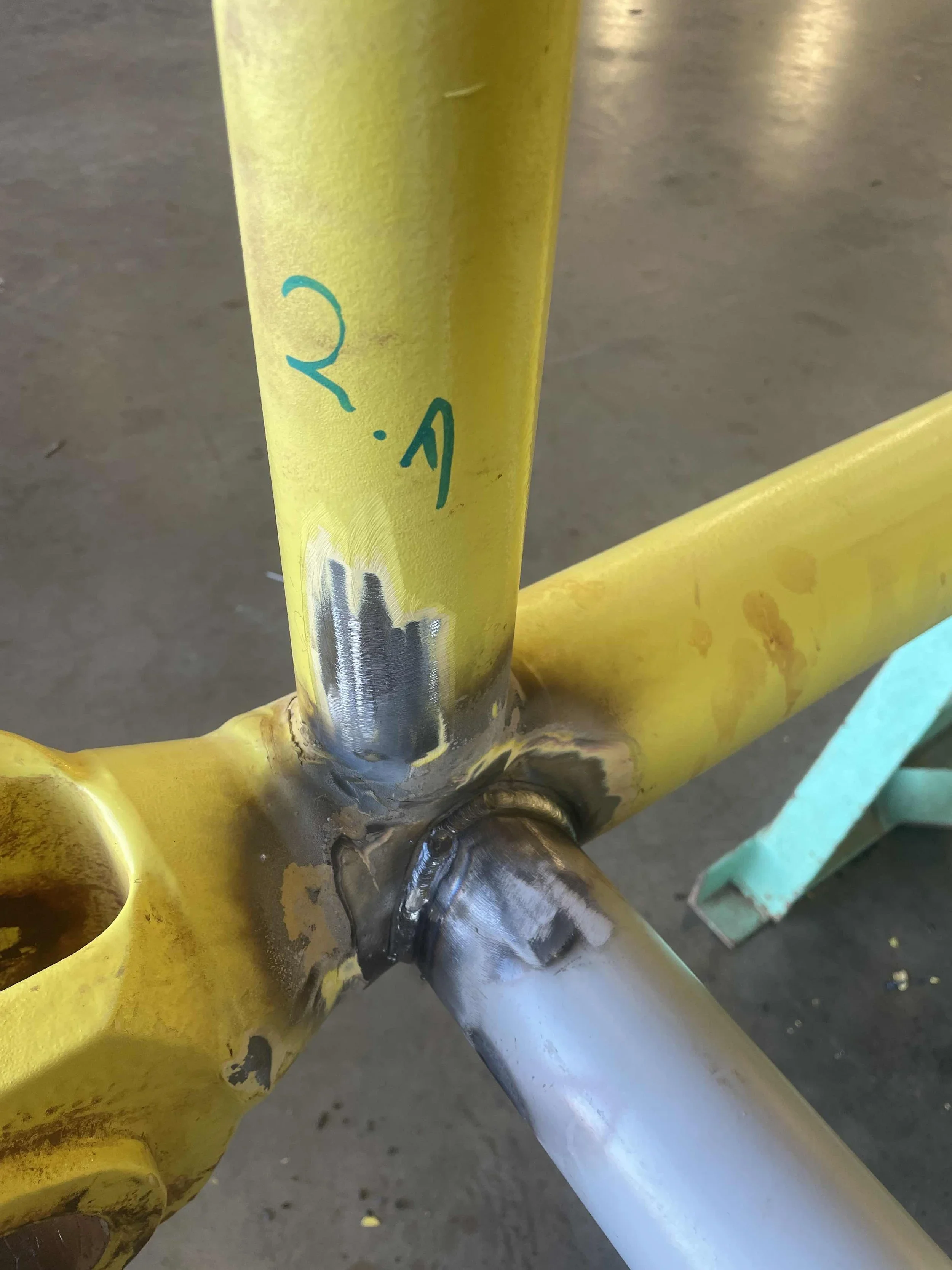 Close-up of a yellow metal bike frame with a welded joint and some handwritten markings in blue ink.
