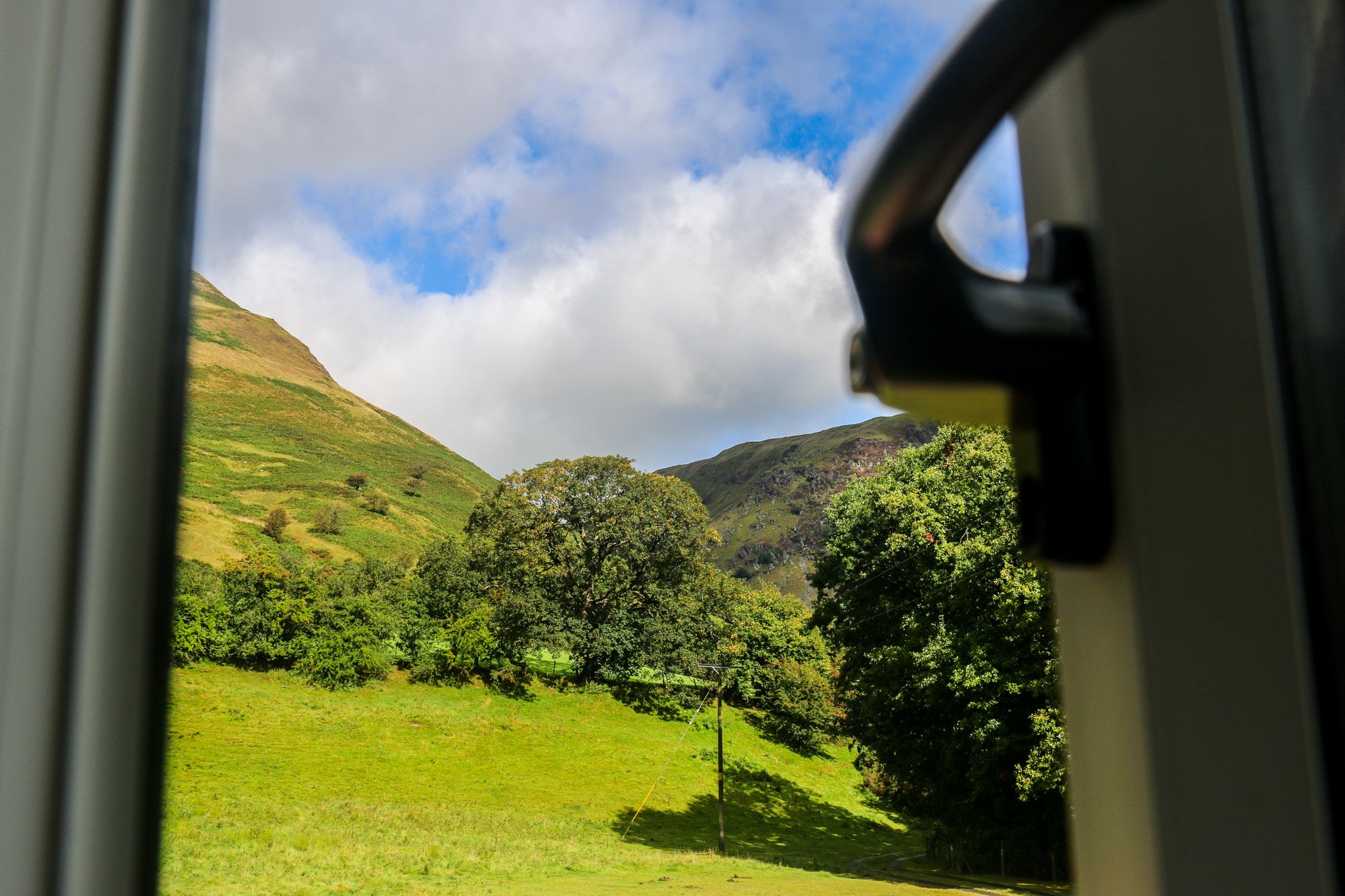 Landscape view of green hills of mid Wales, trees, and a partly cloudy sky, seen through a window with a handle.