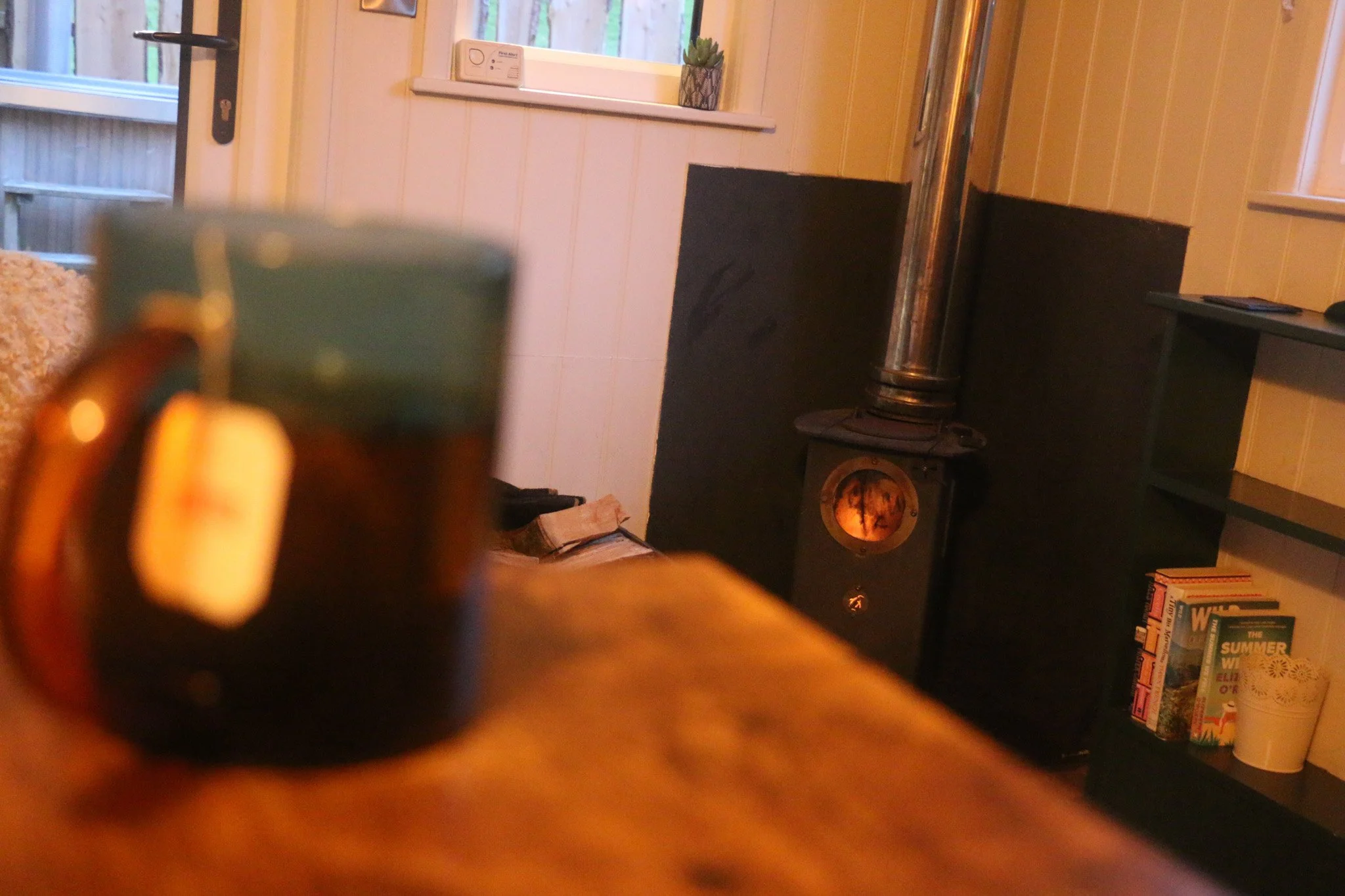 Blurry photo of a Welsh coffee mug on a wooden table with a roaring fire in a wood burner and bookshelf in the background