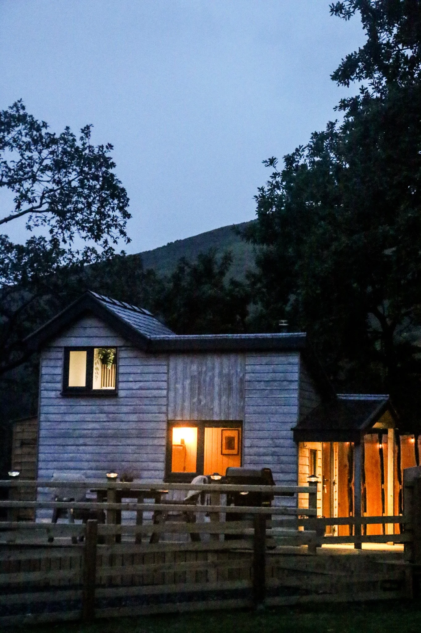 A cabin at dusk with ambience outdoor lighting on, framed by trees and Eryri (Snowdonia) mountain in the background.