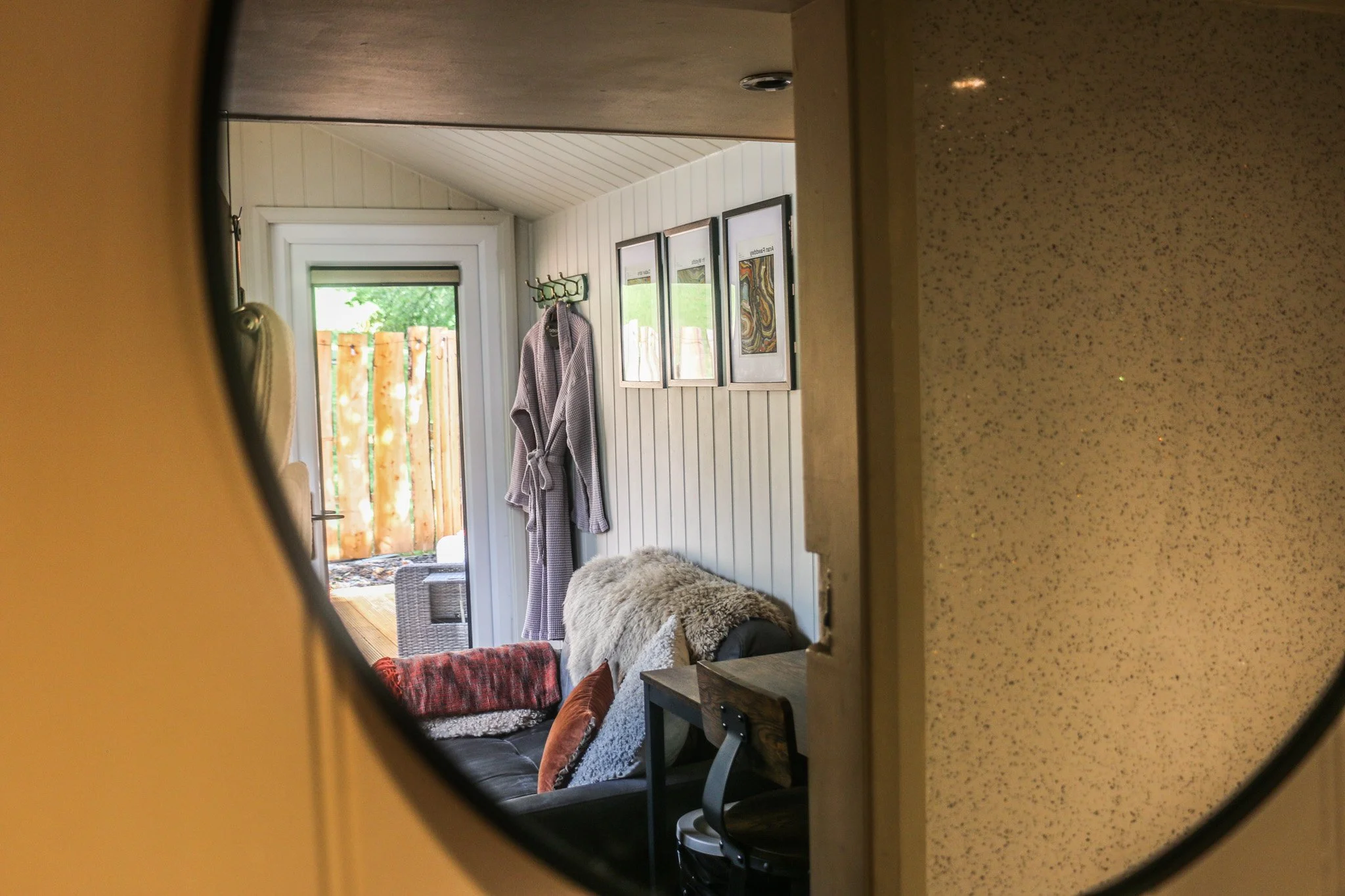 Living room viewed through a mirror, with a comfy couch, pillows, cosy blanket, and Eryri (Snowdonia) wall art, and an outdoor patio with wooden fence and wood fired hot tub visible through a glass door.