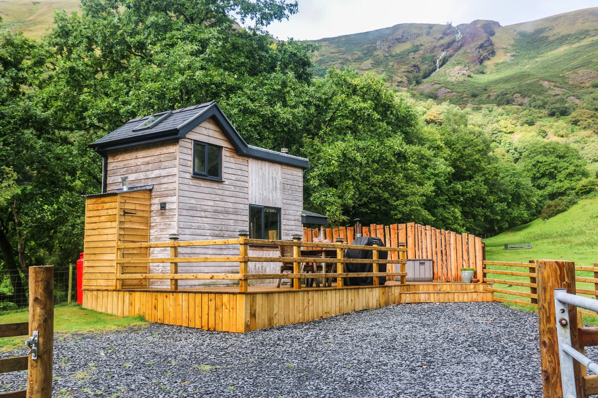 A cute, two-story wooden cabin with black windows, surrounded by a rustic wooden fence, with a mountain, waterfall and trees in the background.