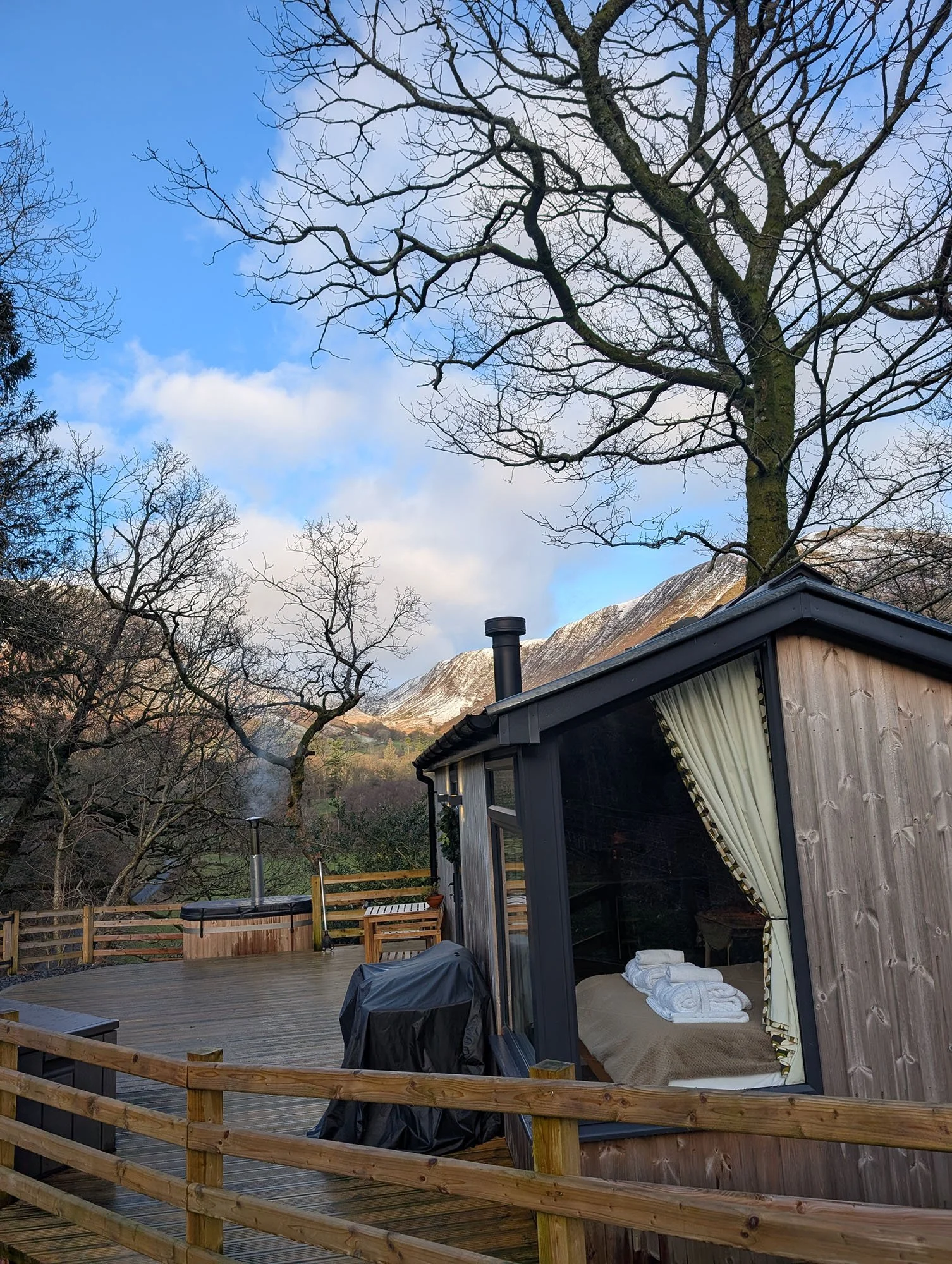 Wooden cabin with large glass window and outdoor deck, towels on cosy bed inside, surrounded by trees and Bwlch y Groes mountains in the background, with partly cloudy beautiful sky.