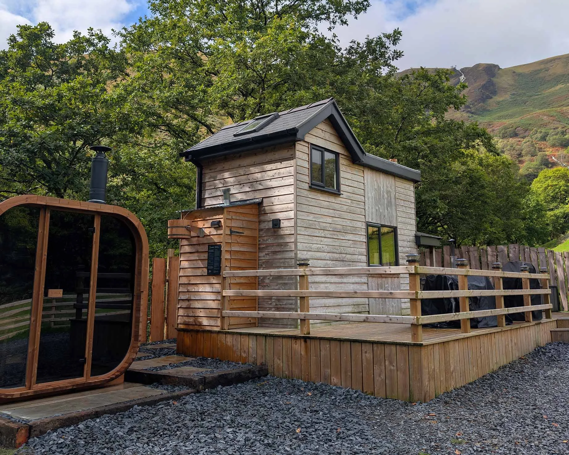Private wooden cabin modern outdoor sauna, enclosed decking area and slate gravel outdoor space. Mountain, waterfall and trees in the background.