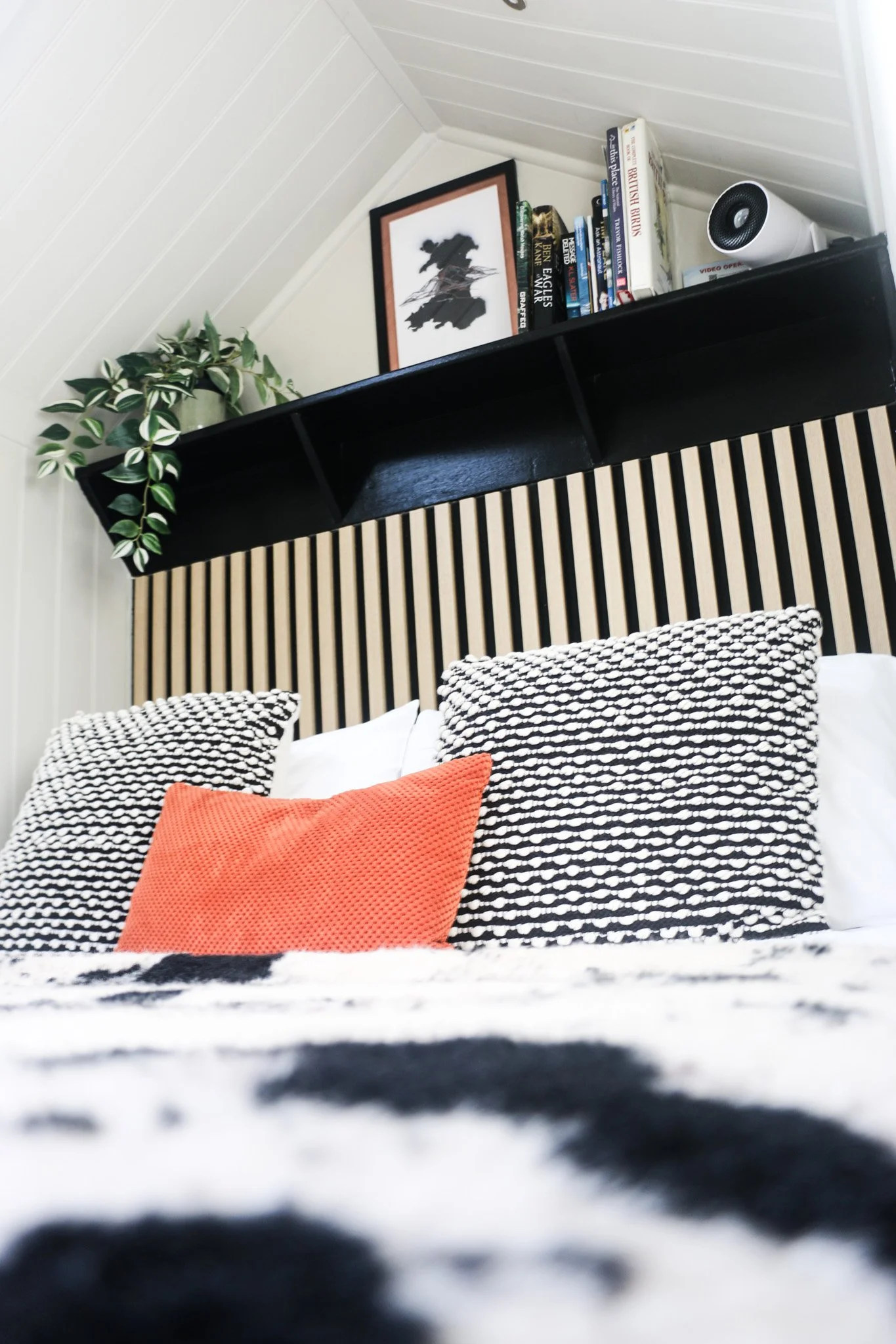 Close-up view of a cosy king sized bed with patterned pillows. The headboard has vertical black and wood slats, with a black shelf above holding ficton and nature books, a picture frame of Wales, a potted plant, and a SMART projector.