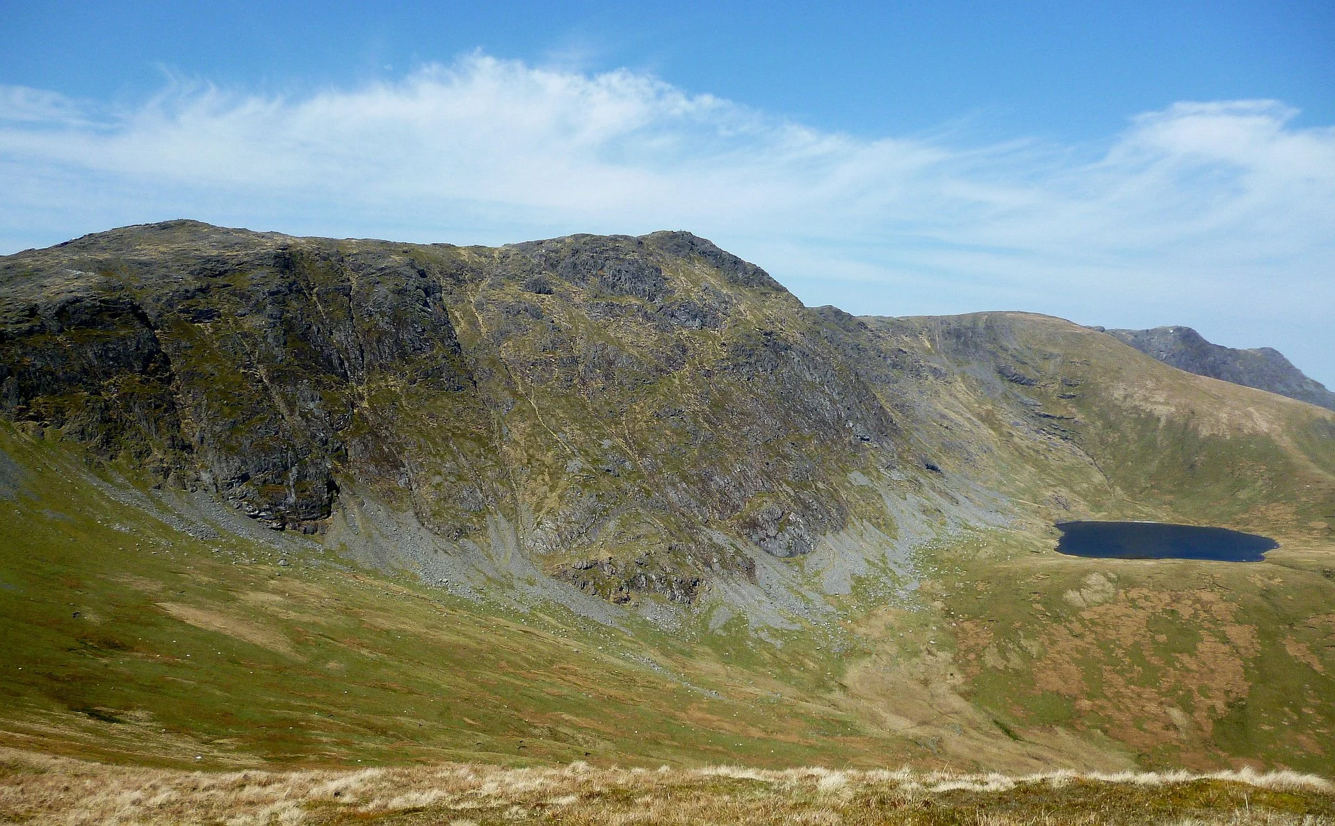A scenic landscape of Aran Fawddwy featuring a large rocky hill with patches of green grass, Creiglyn Ddyfi lake to the right, and a partly cloudy blue sky overhead.
