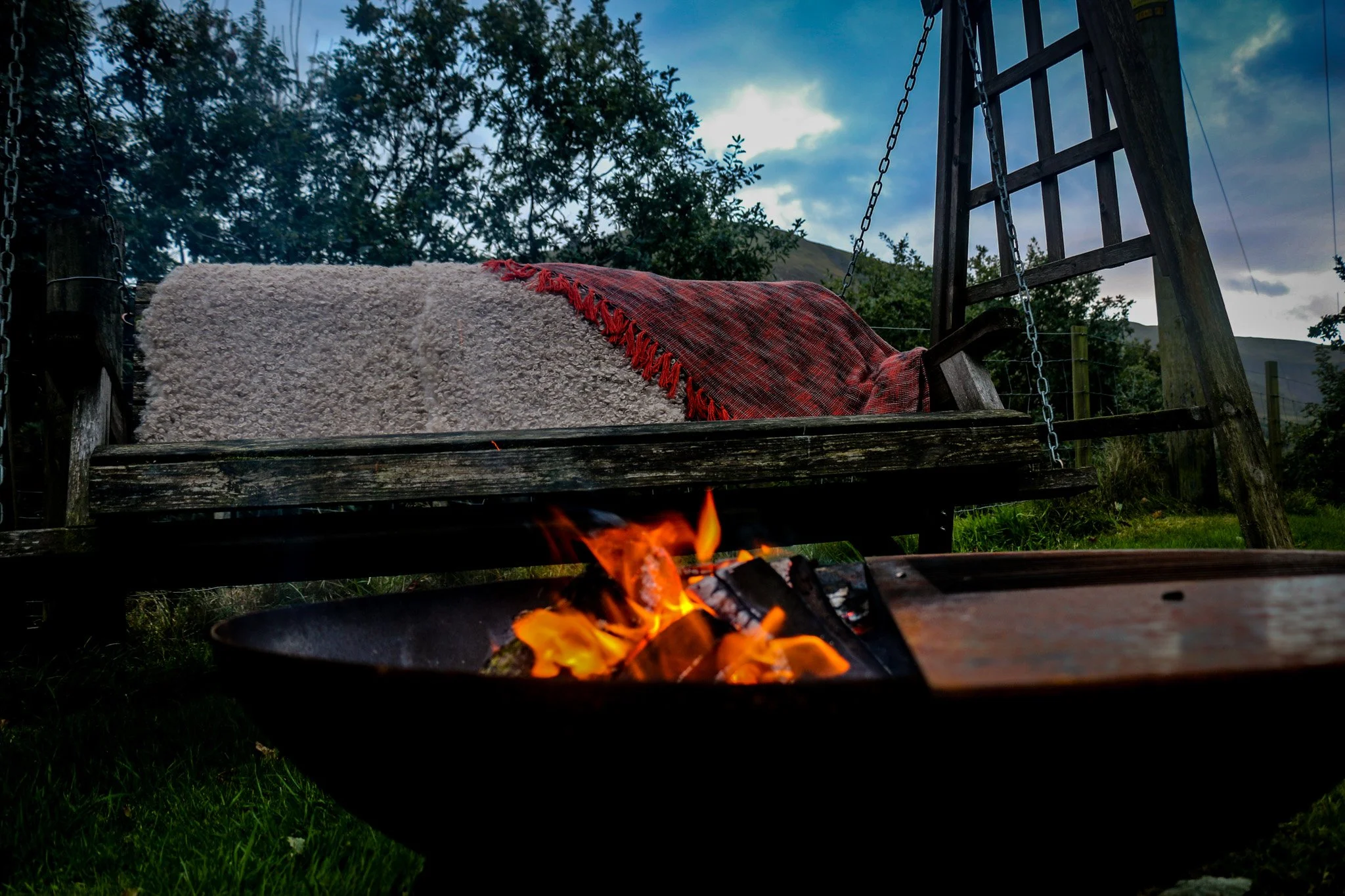A rustic outdoor swing with a orange blanket, set against a backdrop of trees and mountains of Eryri (Snowdonia), with a fire pit burning in the foreground.