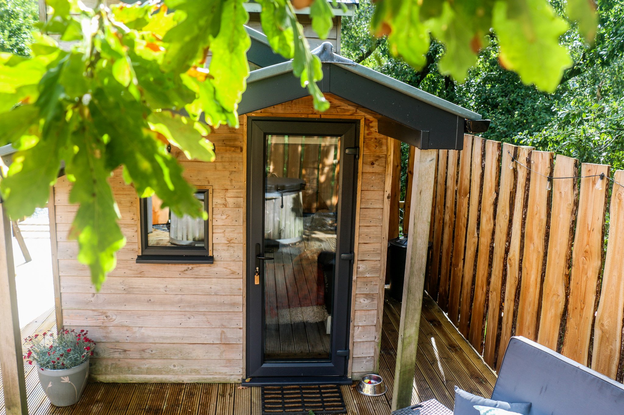 A small wooden cabin with a glass black door and window, surrounded by a wooden deck and fence. There is a pot with flowers and an outdoor sofa to relax and enjoy the view in the foreground.