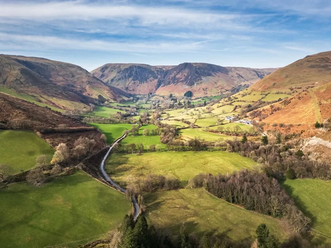 A lush green valley surrounded by rolling hills with a winding road through it, under a partly cloudy sky.