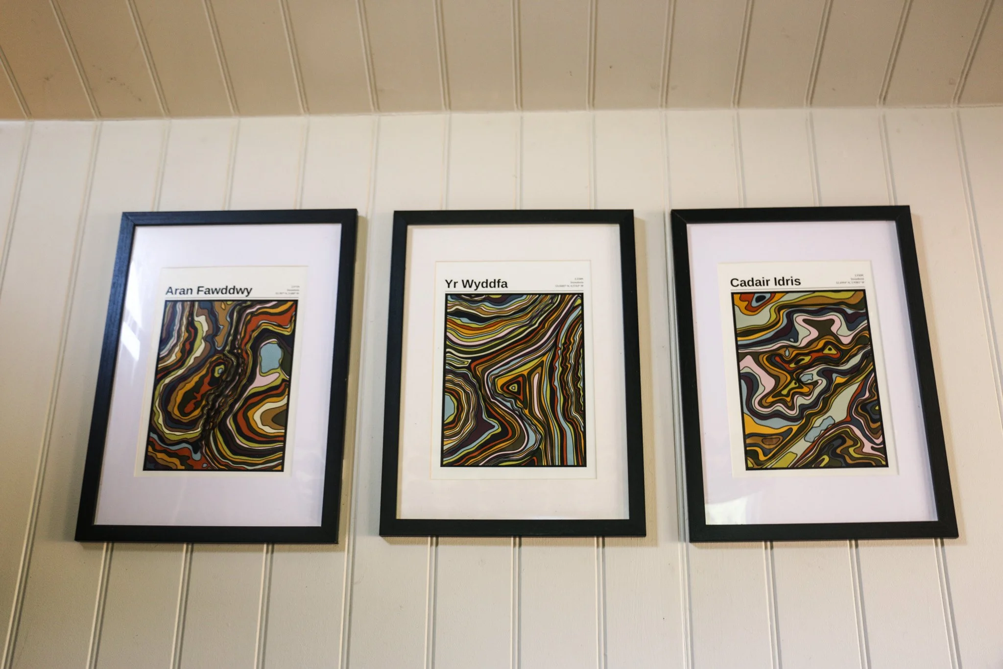 Three framed abstract colorful artwork pieces on white wooden wall with labels in Welsh above each piece of Aran Fawddwy, Wyddfa and Cadair Idris (Snowdonia/Eryri mountains).
