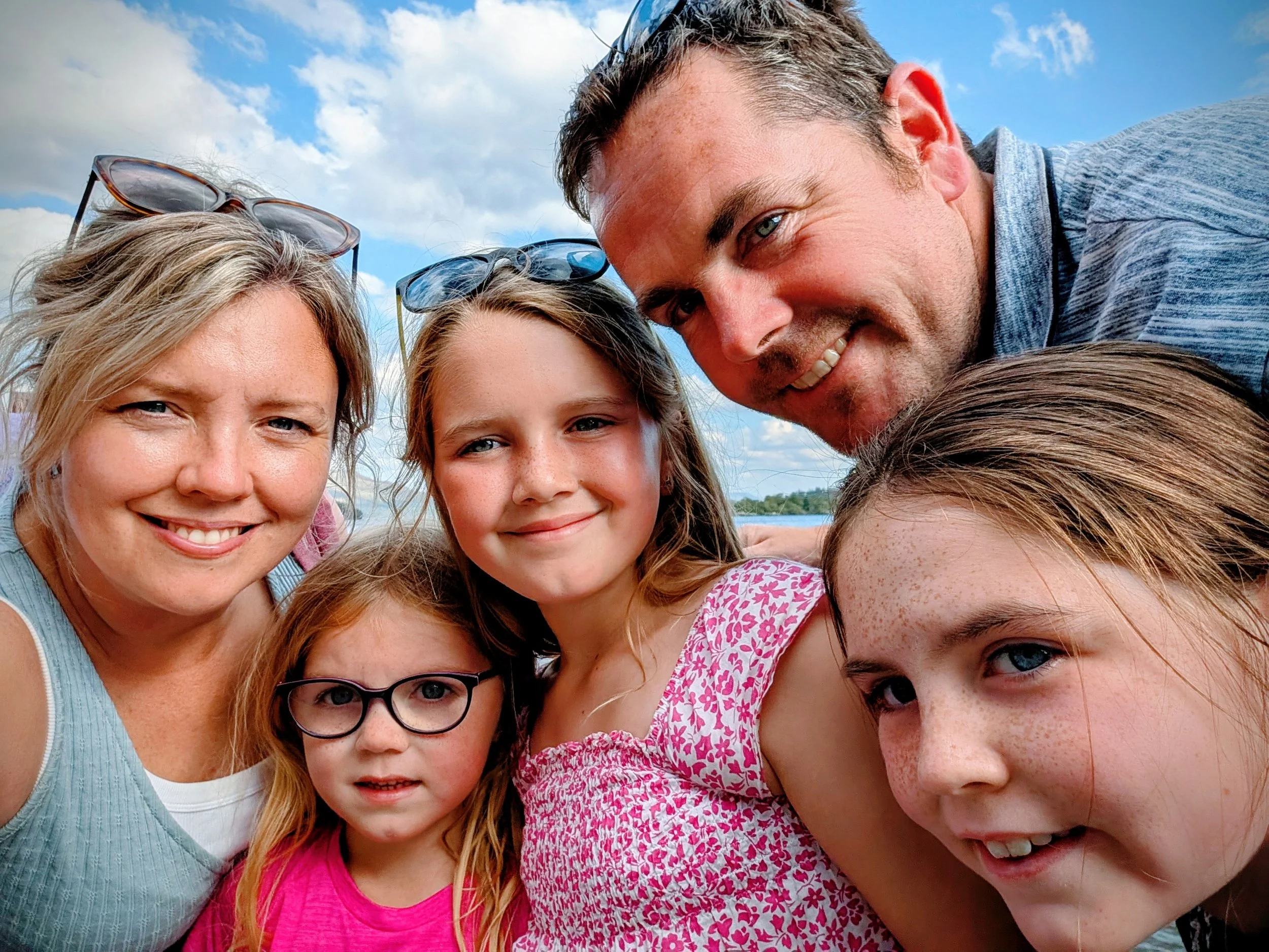 A family of five taking a selfie outdoors on a sunny day, with a blue sky and some clouds in the background.