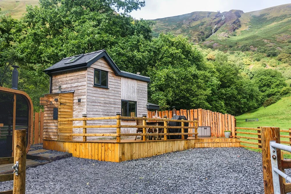 Modern timber holiday cabin with sauna and decking area within a fenced enclosure and slate gravel, set within mature oak trees in autumnal colours, set in the hills.
