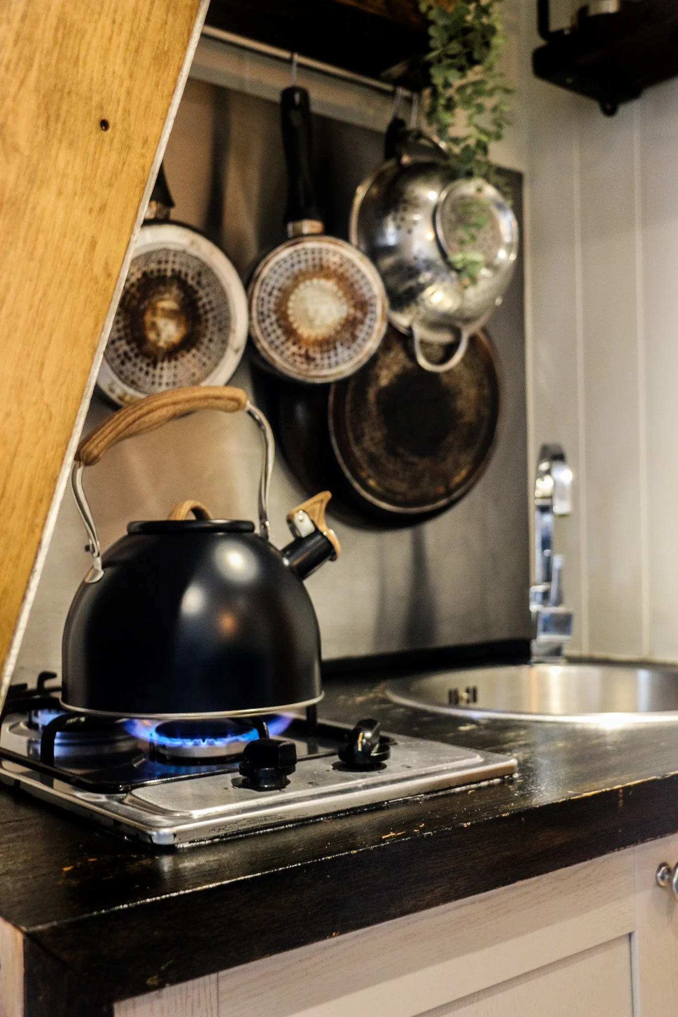 A kitchen stove with a black tea kettle on the gas hobs, with pots and pans hanging on the wall behind, and a sink with a faucet to the right. Perfect for cooking romantic dinners for two.