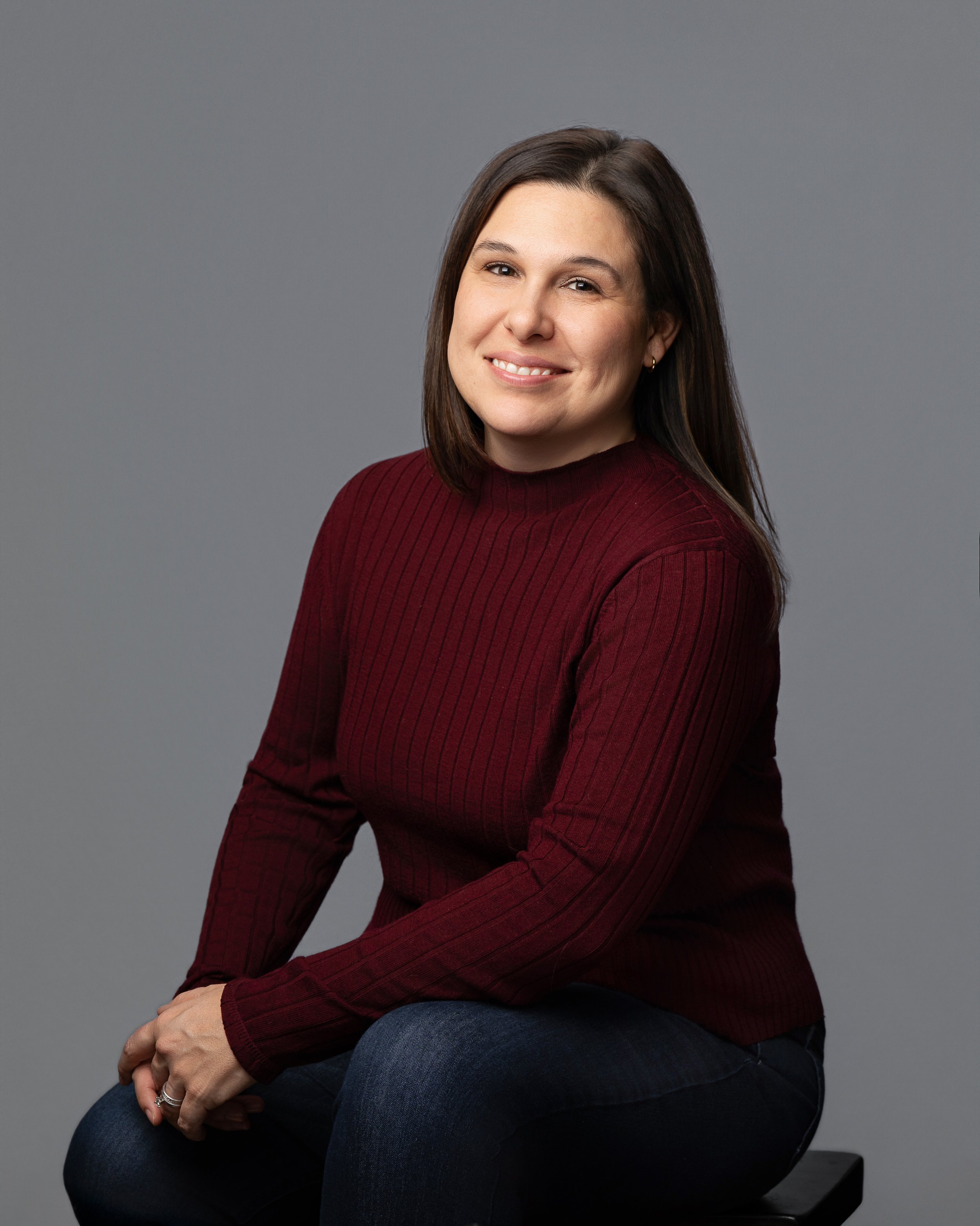 Headshot of a young woman with dark brown hair, smiling, wearing a maroon sweater and jeans, sitting against a gray background.