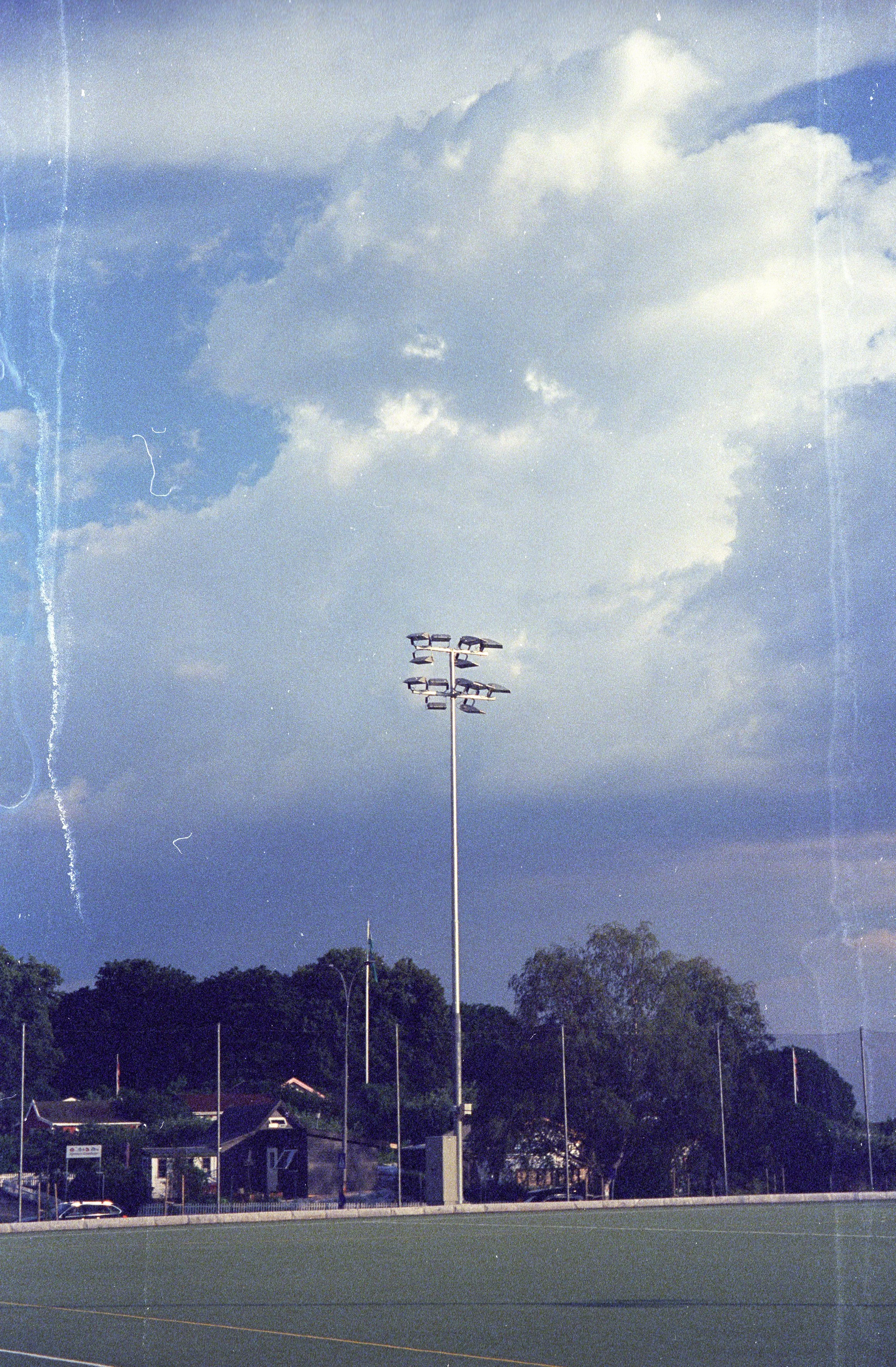 An outdoor sports field with a tall stadium light pole, trees, and small buildings in the background under a cloudy sky.