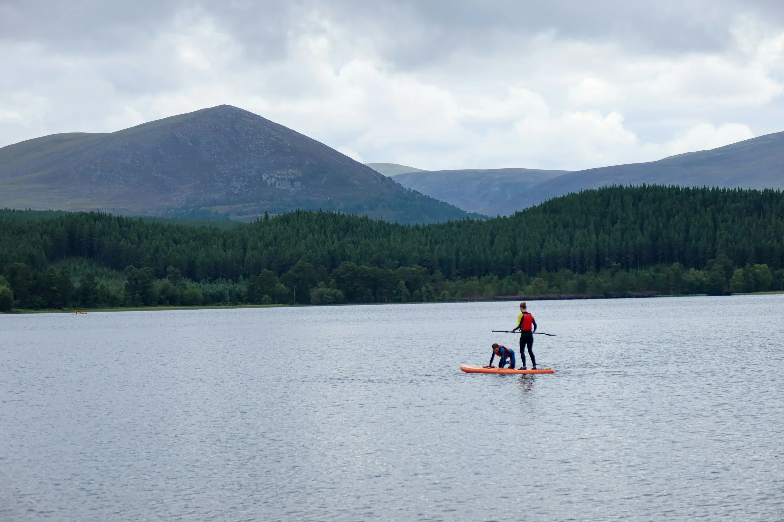 Two people stand on a paddleboard on a calm lake, with a backdrop of mountains and dense green trees, under a partly cloudy sky.