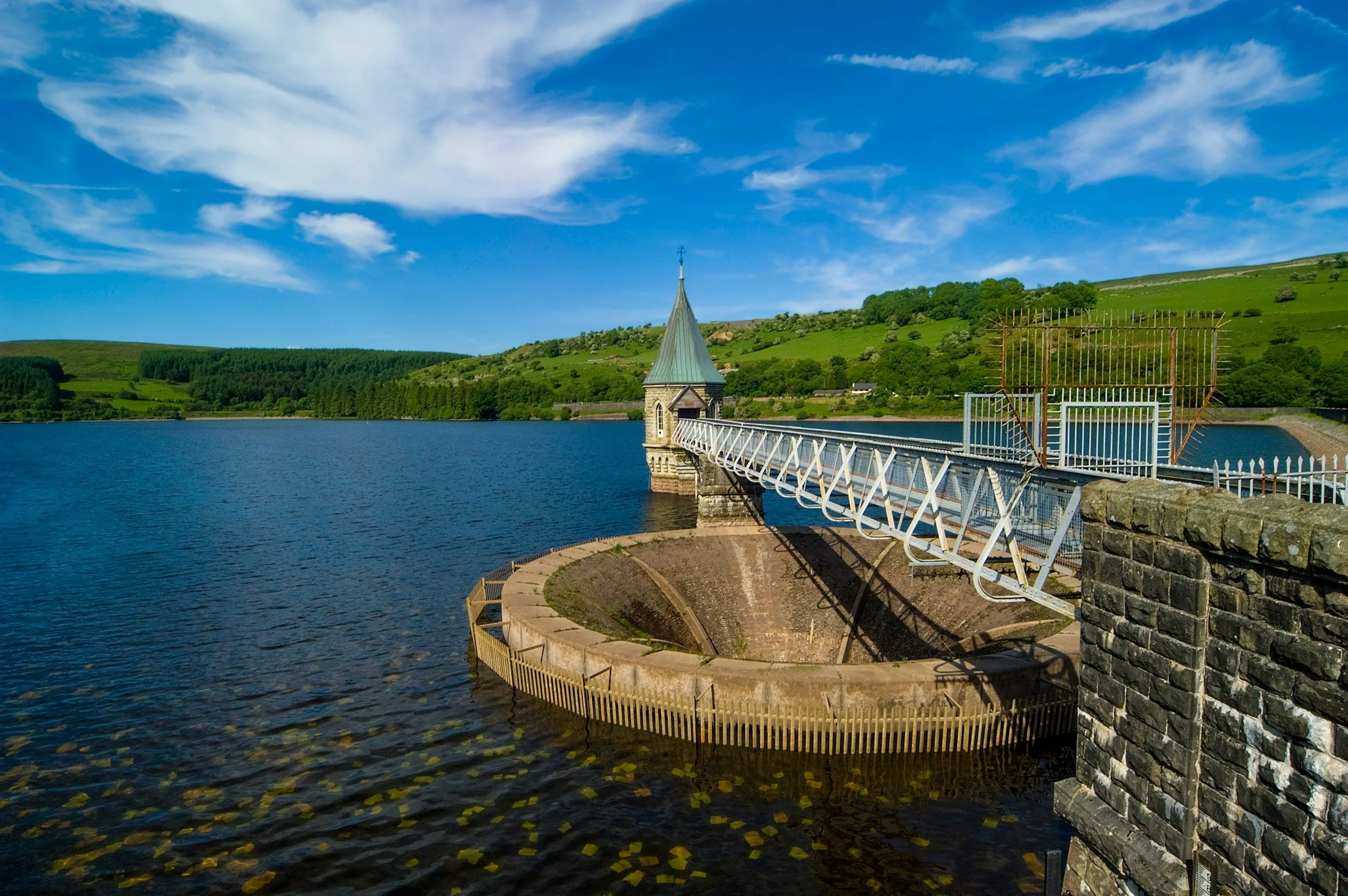 A scenic view of a dam with a small tower and a bridge extending over a body of water, with green hills and trees in the background and a blue sky with some clouds.